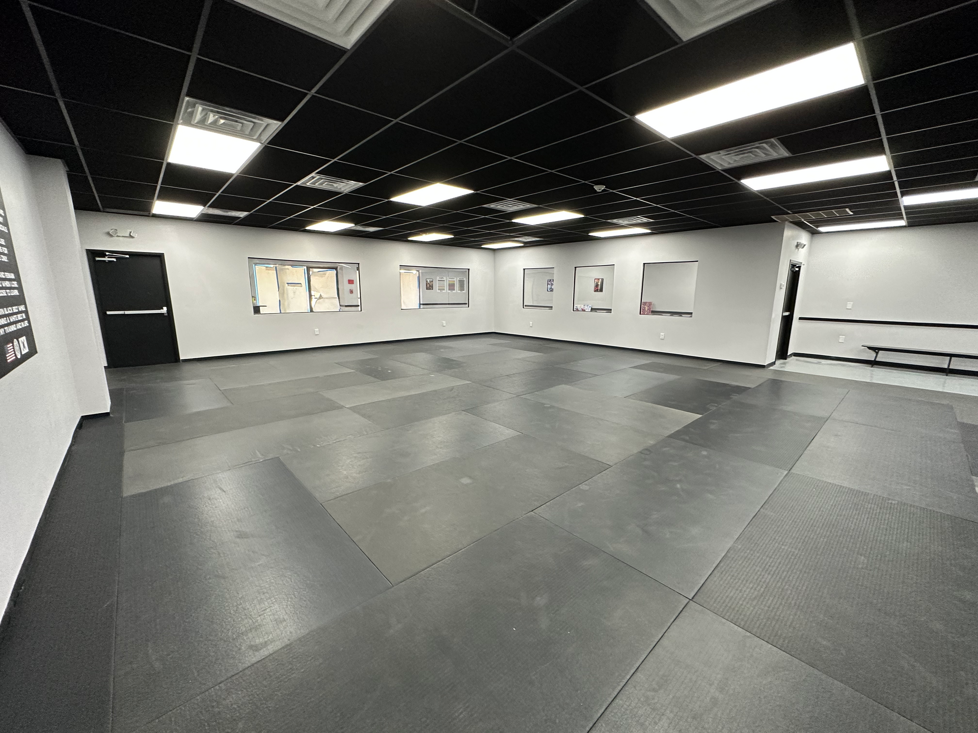 A wide, empty fitness studio with black matted flooring, white walls, and a black drop ceiling with rectangular lights.