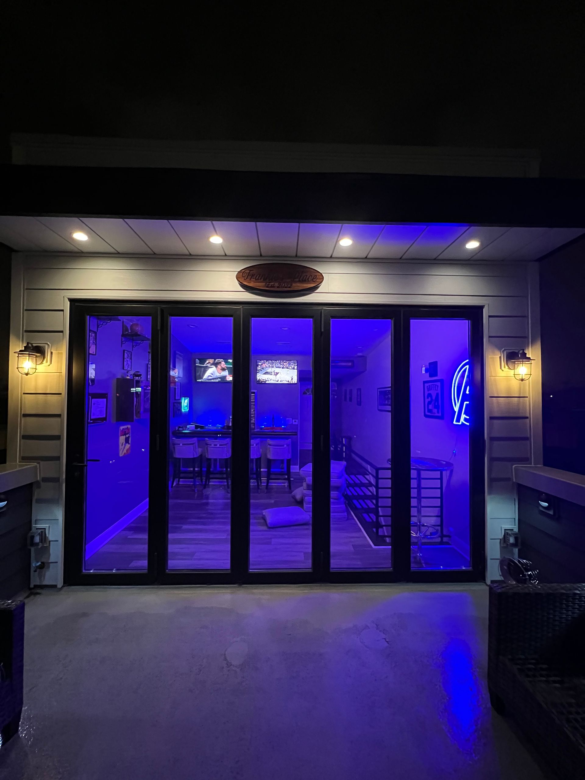 A dimly lit patio at night, looking into a modern indoor lounge with floor-to-ceiling glass doors and vibrant blue lighting.