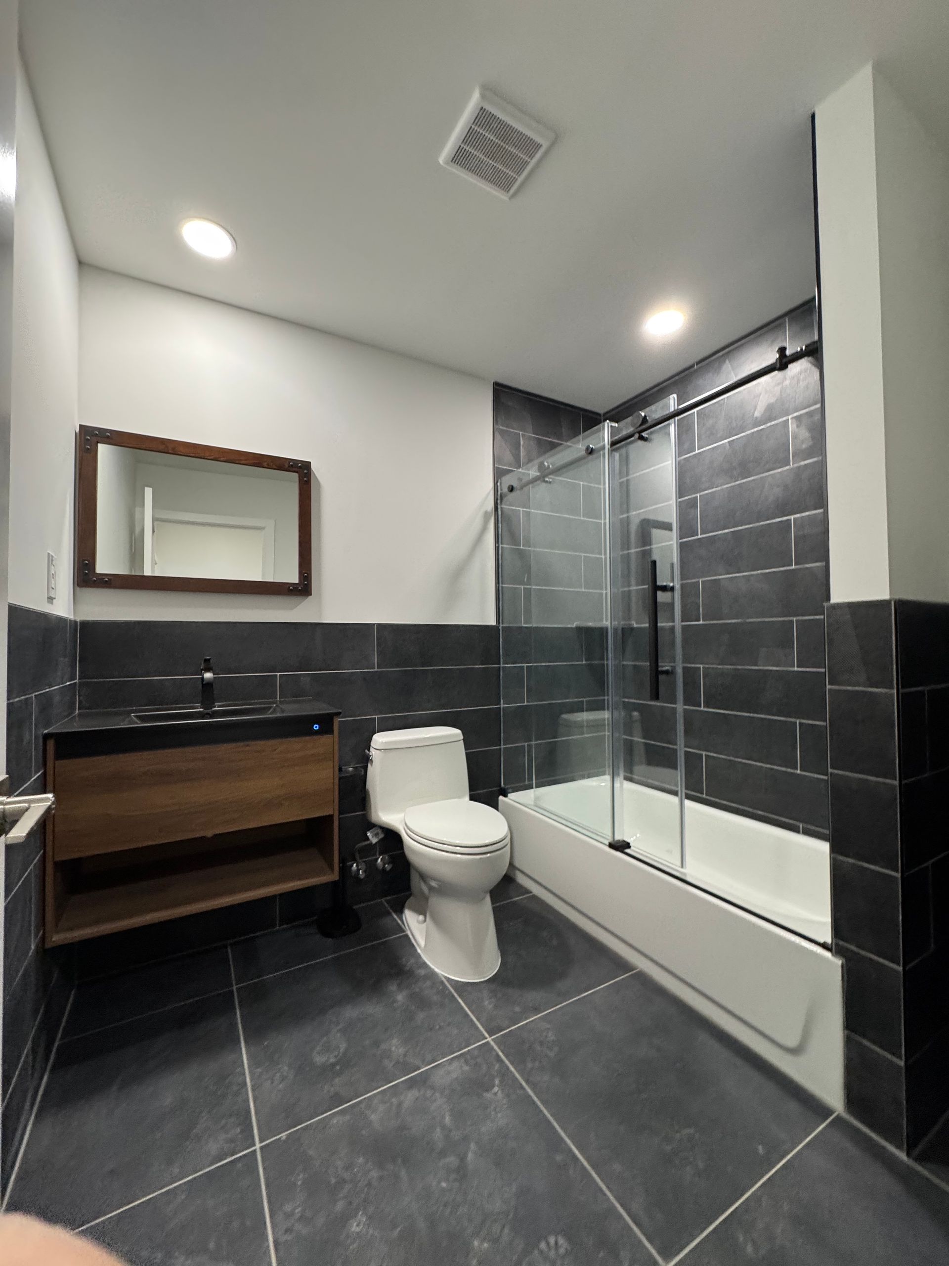 A modern bathroom with dark grey tiles, a floating wood vanity, a white toilet, and a glass-enclosed bathtub.