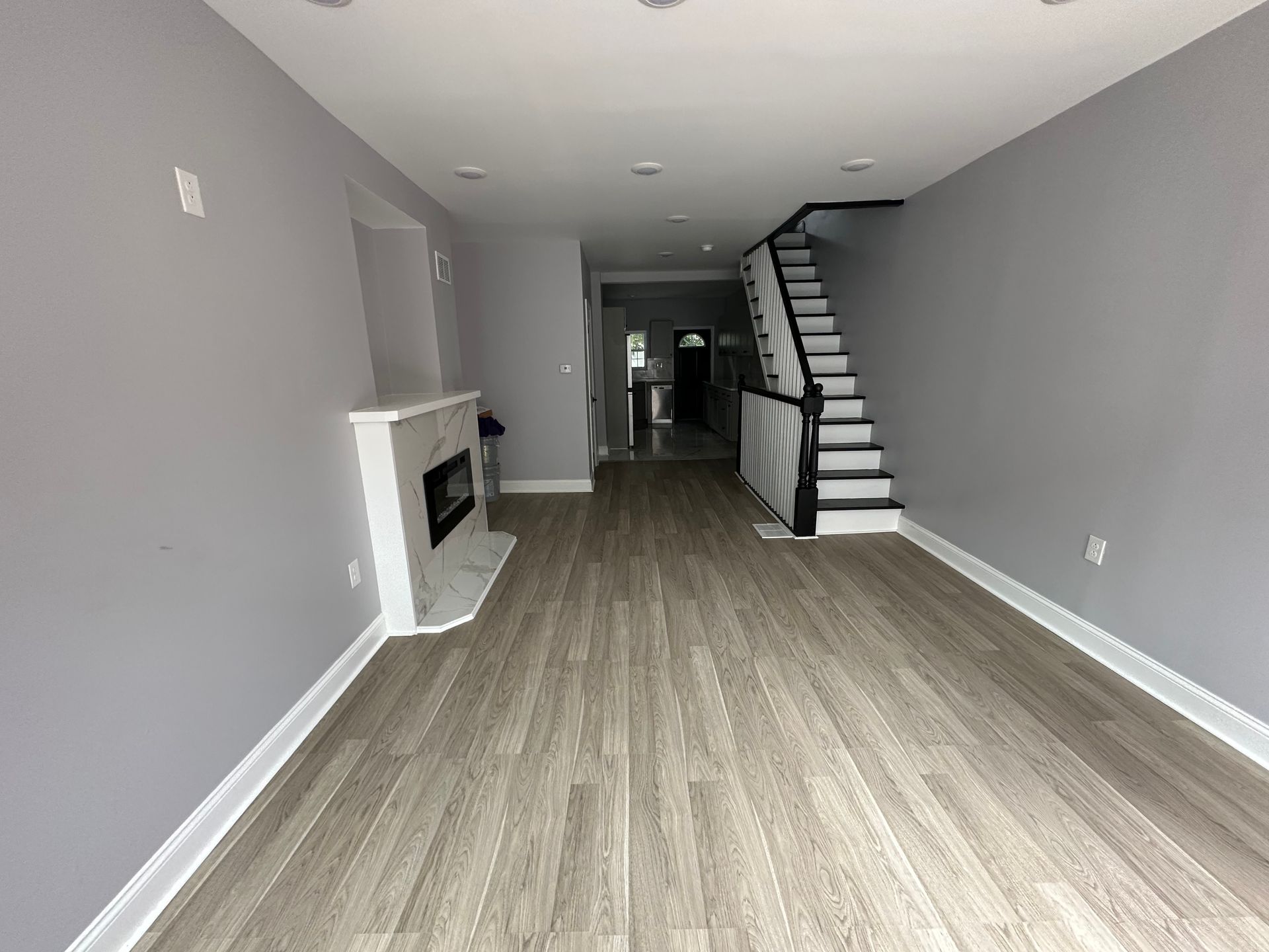 A modern, empty living room with gray walls, wood-look flooring, a white marble fireplace, and a staircase.