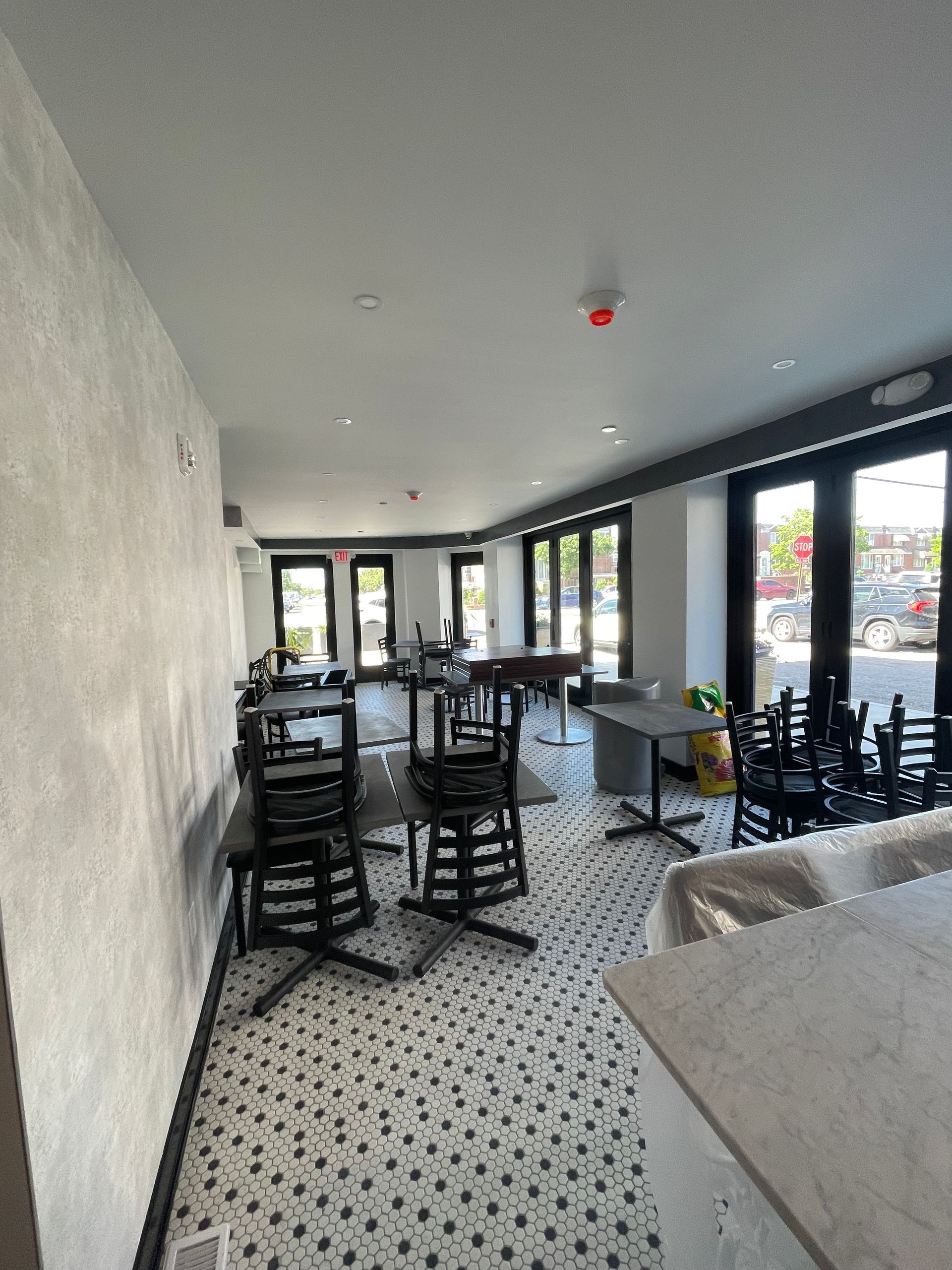 Interior of a cafe with tables and stacked chairs on a patterned tiled floor, featuring large glass doors and grey walls.