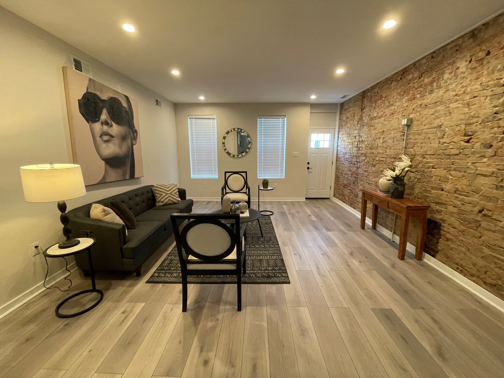 A living room features light hardwood floors, a dark sofa, two chairs, a wooden console table, and an exposed brick wall.