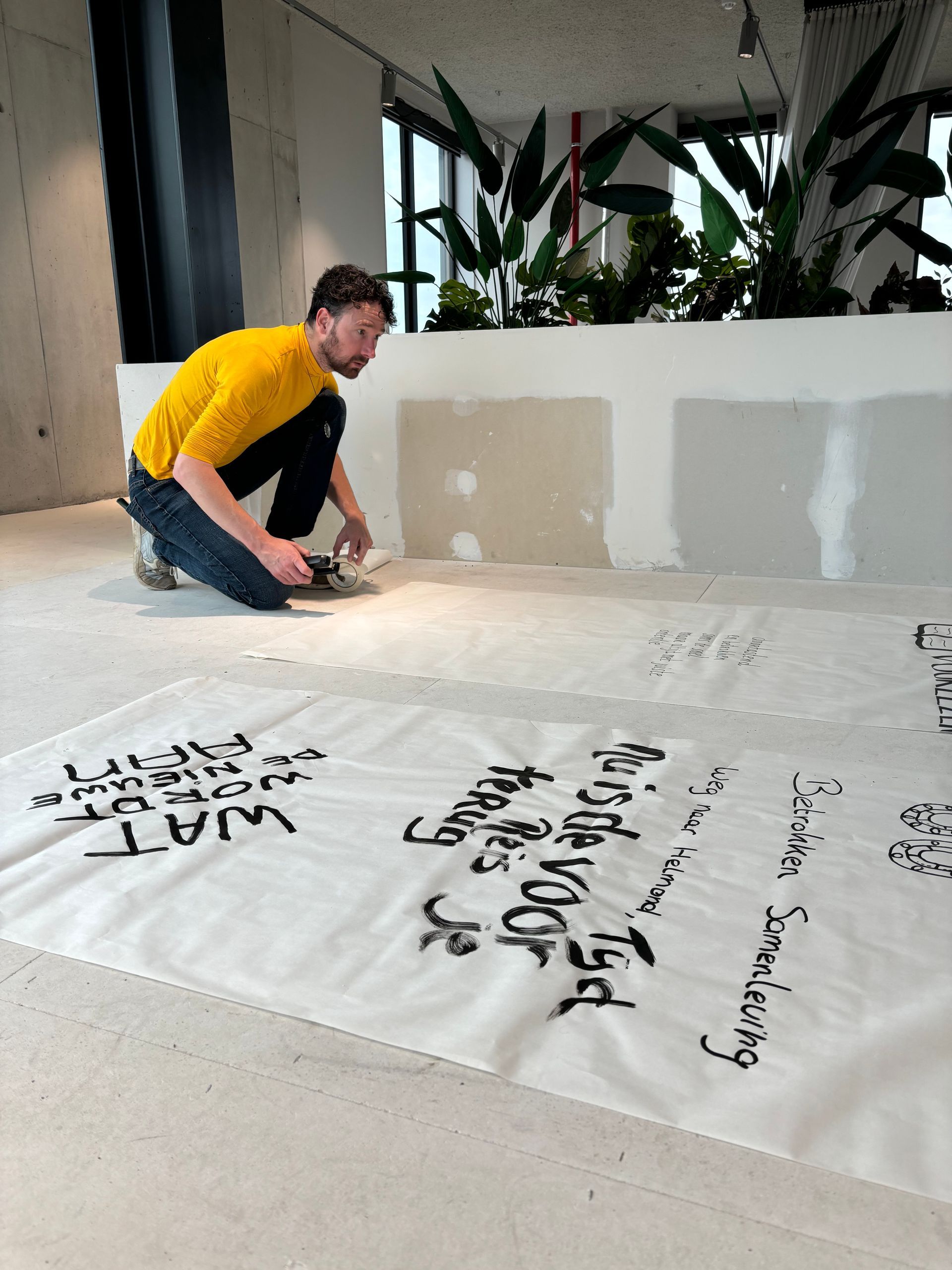 Een man spuit tekst op een wit laken op de vloer, vlakbij een witte muur en planten. Hij draagt ​​een geel shirt en een blauwe spijkerbroek.