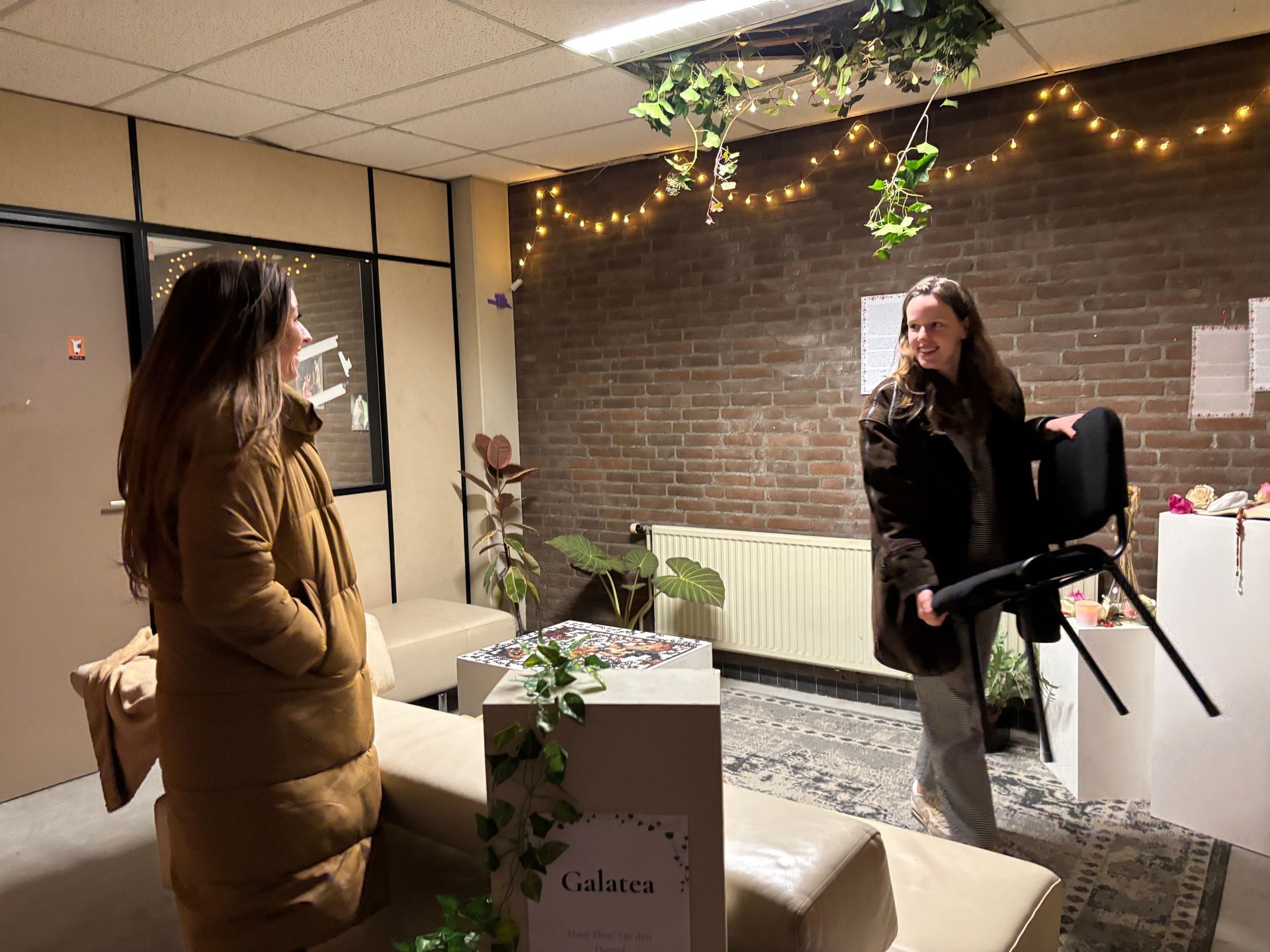 Twee mensen in een kamer versierd met planten en lampen; de een draagt ​​een stoel, de ander kijkt toe.