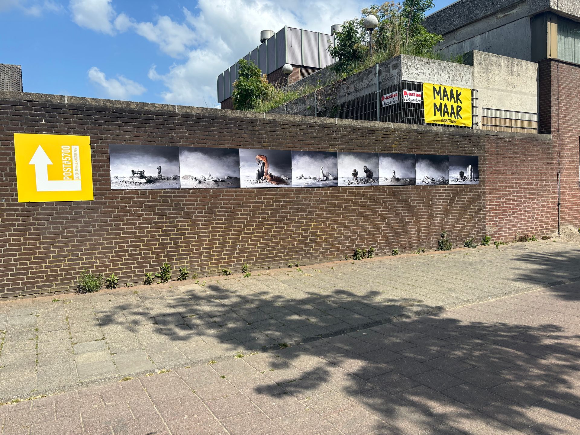 Bakstenen muur met een reeks zwart-witfoto's. Gele borden met pijlen en tekst. Zonnige buitenomgeving.