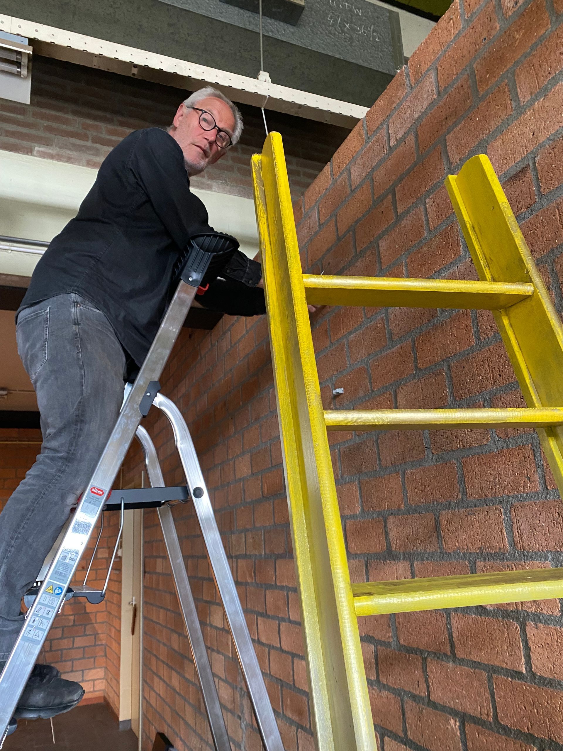 Man op een ladder bij een bakstenen muur, met een gereedschap in zijn hand. Gele en zilveren ladder, binnenomgeving.
