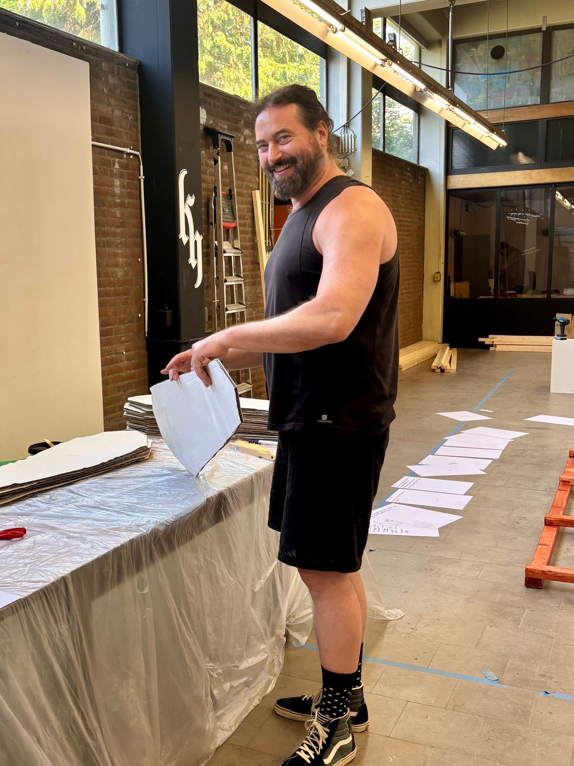 Een man in een zwart hemdje en korte broek glimlacht terwijl hij papieren vasthoudt bij een afgedekte tafel in een werkplaats.