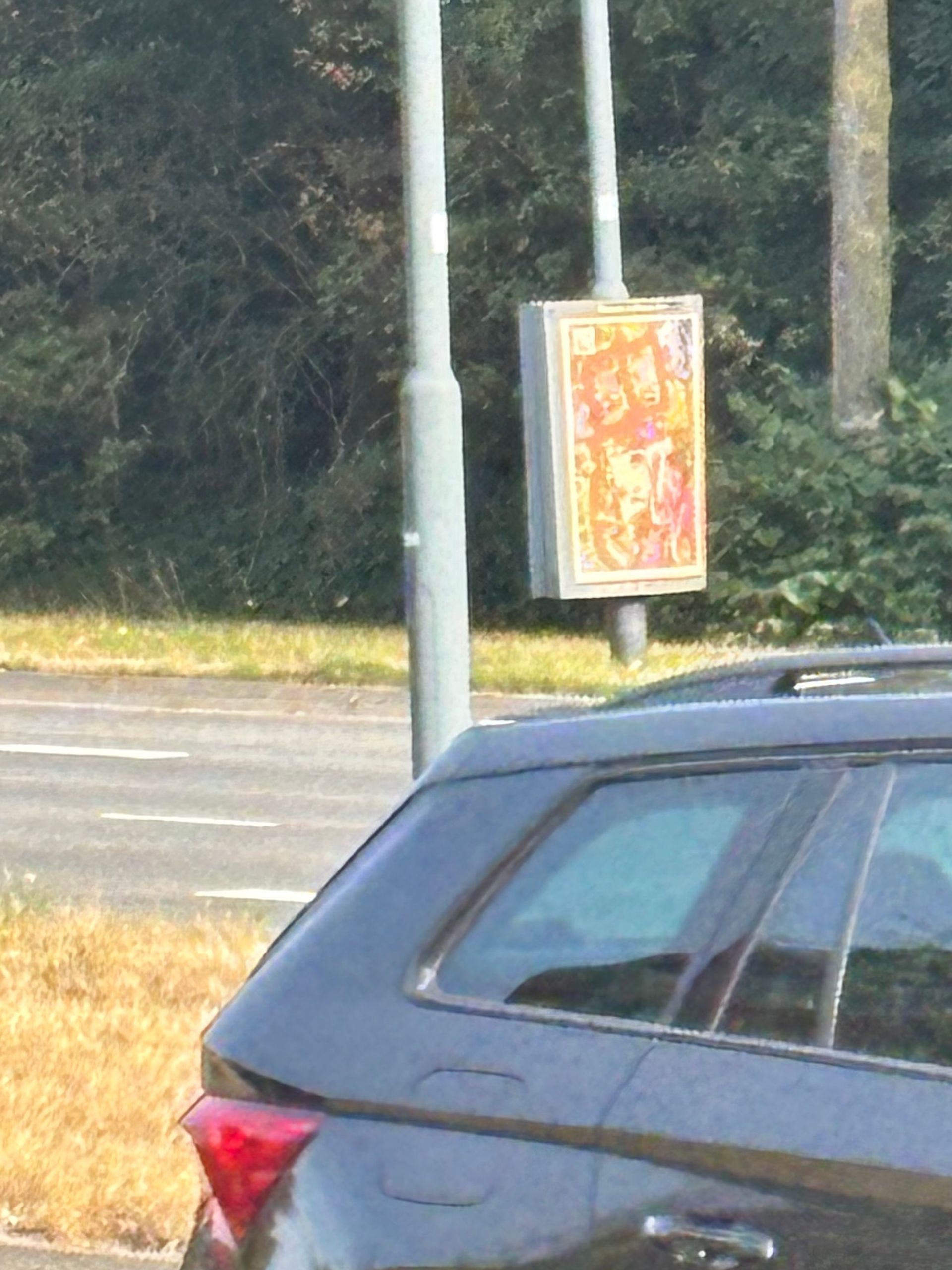 Een zwarte auto staat geparkeerd naast een lantaarnpaal met een kleurrijke poster. Op de achtergrond zijn de weg en bomen te zien.