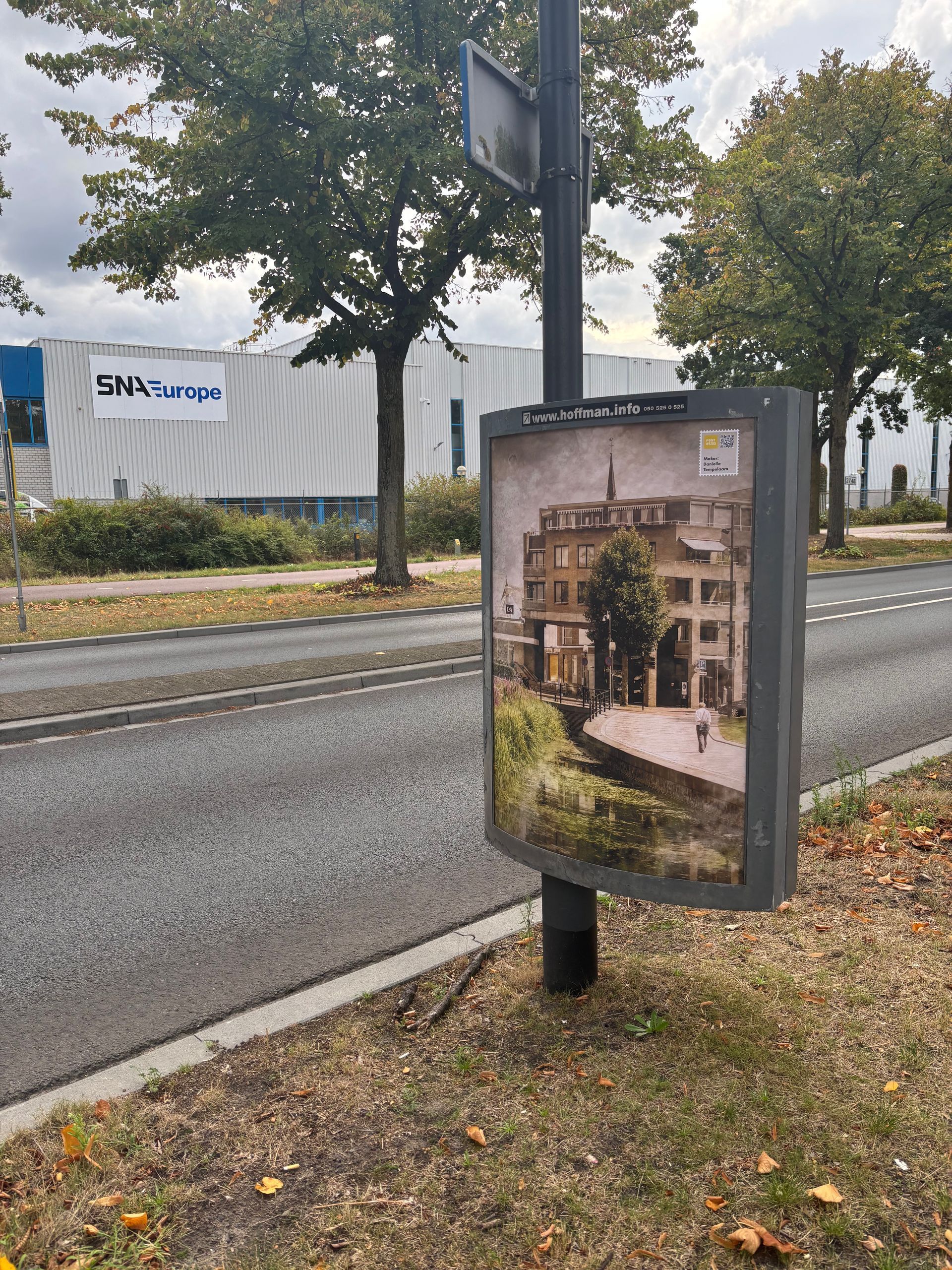 Een reclamebord op een paal toont een tafereel uit een oud stadje naast een weg, met een gebouw op de achtergrond.