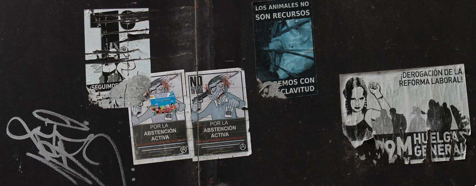 Muur vol gescheurde posters, sommige met tekst. Op één poster staat een persoon afgebeeld. Er is graffiti op de muur.