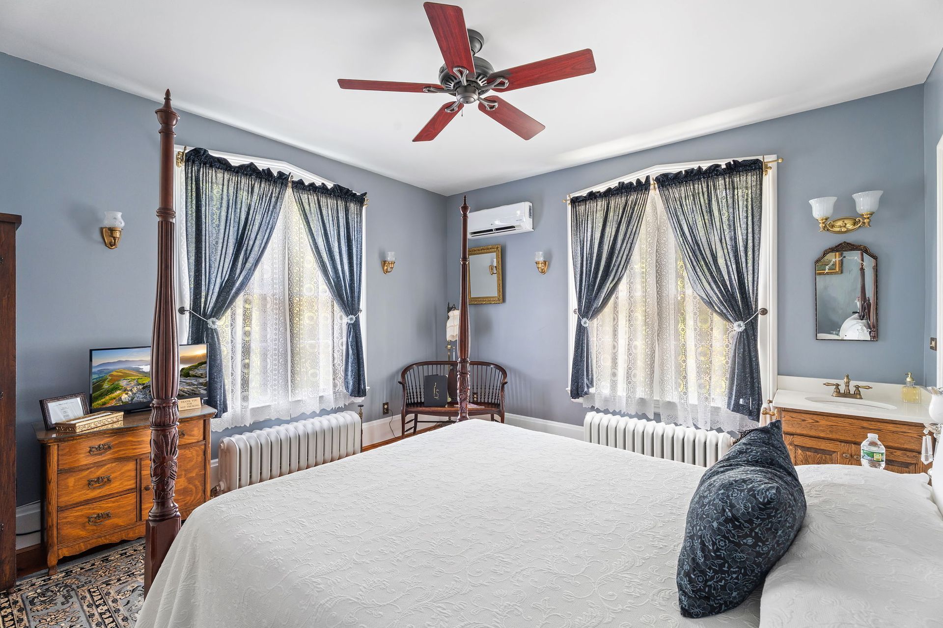 Bright bedroom with twin windows, blue walls, white bed, wood furniture, and red ceiling fan.