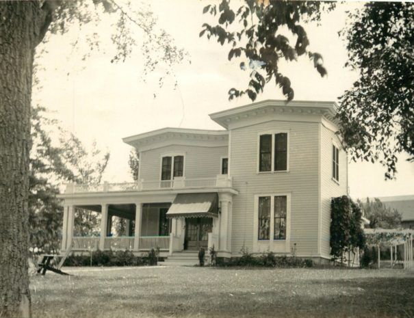 Sepia-toned historic two-story house with porch, framed by trees and a lawn