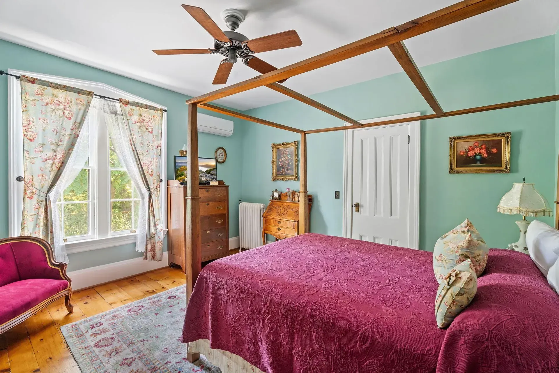 Bright bedroom with four-poster bed, teal walls, pink chair, and large window with floral curtains