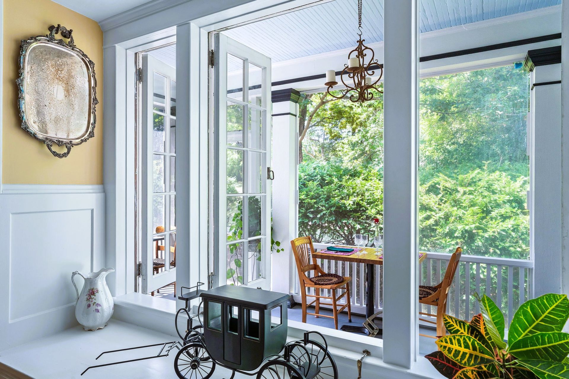 Bright sunroom with open French doors, wicker dining set, and lush green garden view.