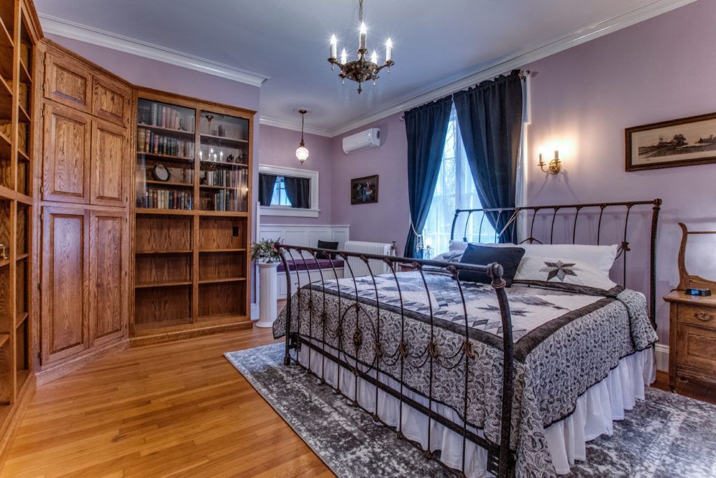 Traditional bedroom with iron bed, wooden floor, built-in shelves, blue curtains, and soft purple walls