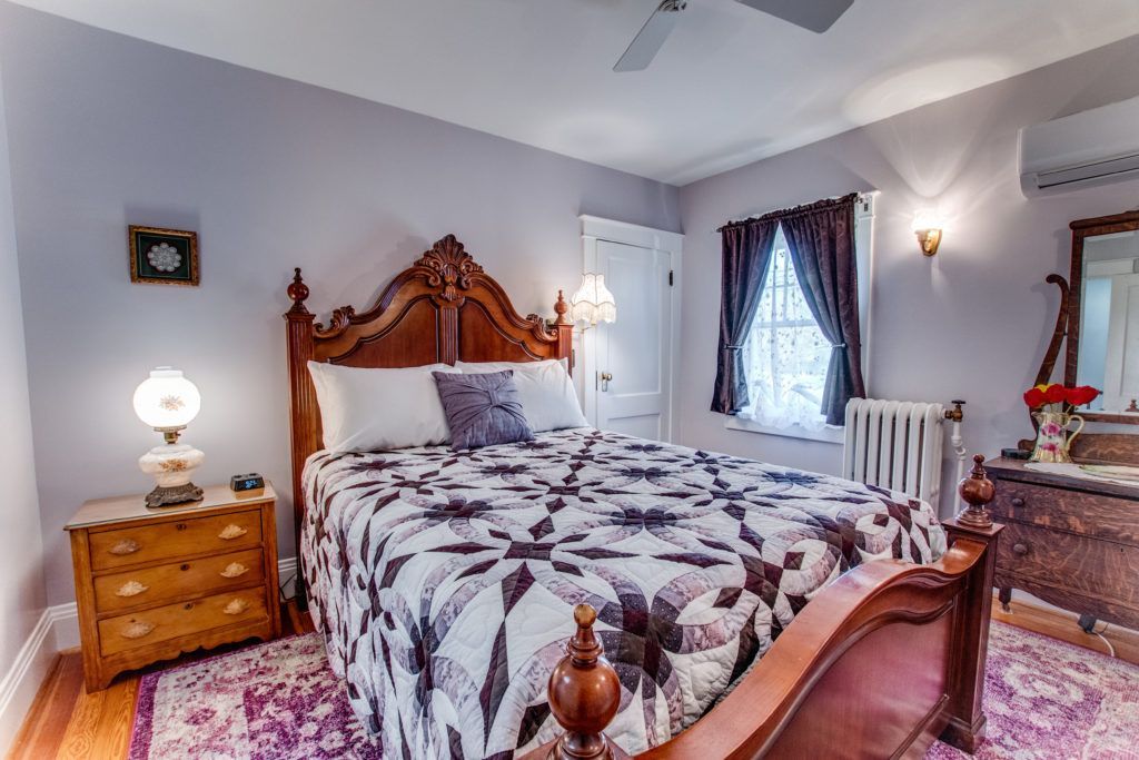 Elegant bedroom with ornate wooden bed, floral bedding, nightstands, window, and dresser in soft gray and purple tones