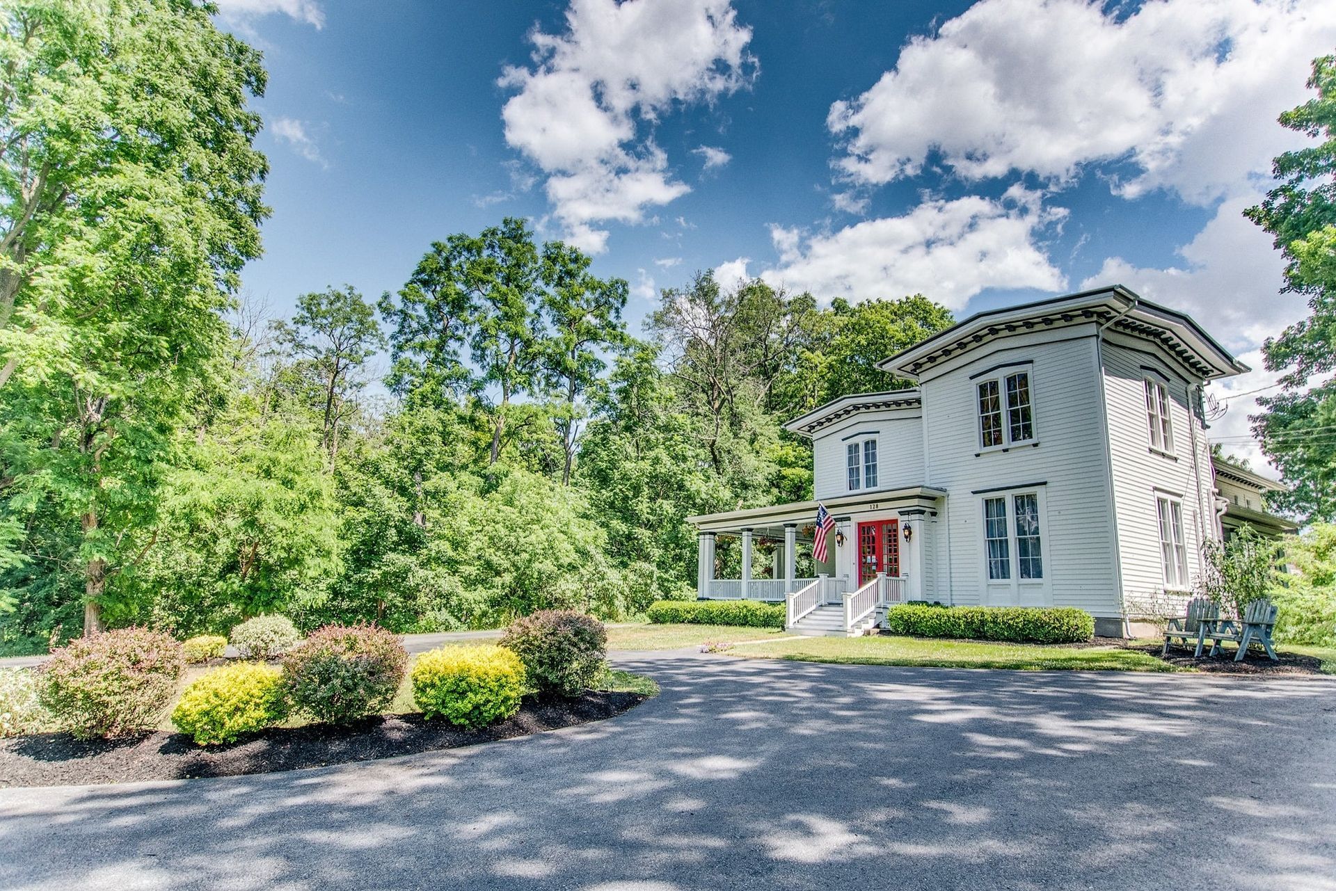 White two-story house with red door, set among trees and shrubs beside a gravel driveway under a blue sky