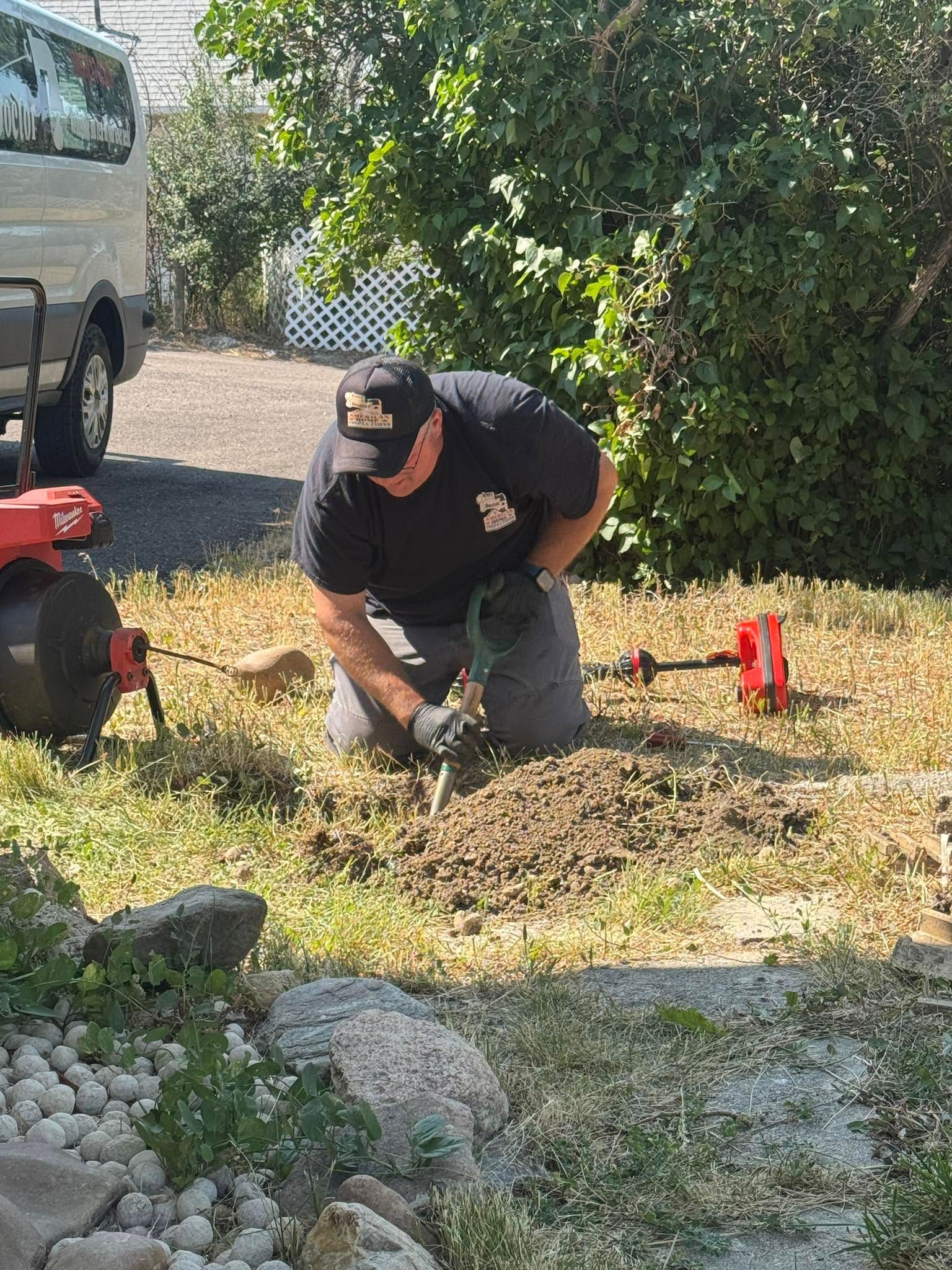 Person kneeling outdoors, using a chainsaw to cut branches beside a truck and red gas can.