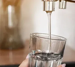 Water pours from a metallic kitchen faucet into a clear glass held by a hand.