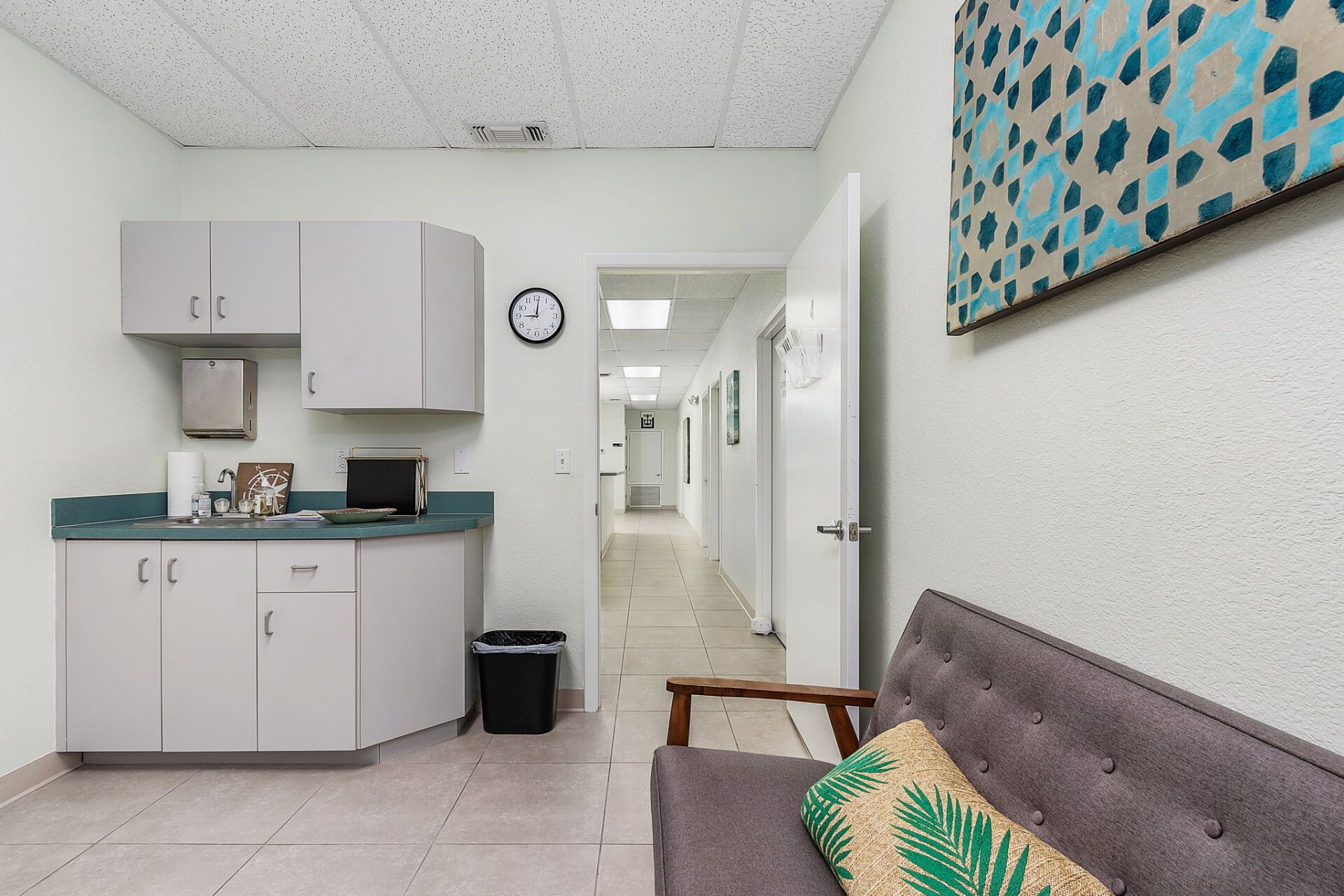 Small clinic waiting area with kitchenette, hallway, wall clock, and a sofa with patterned pillow.