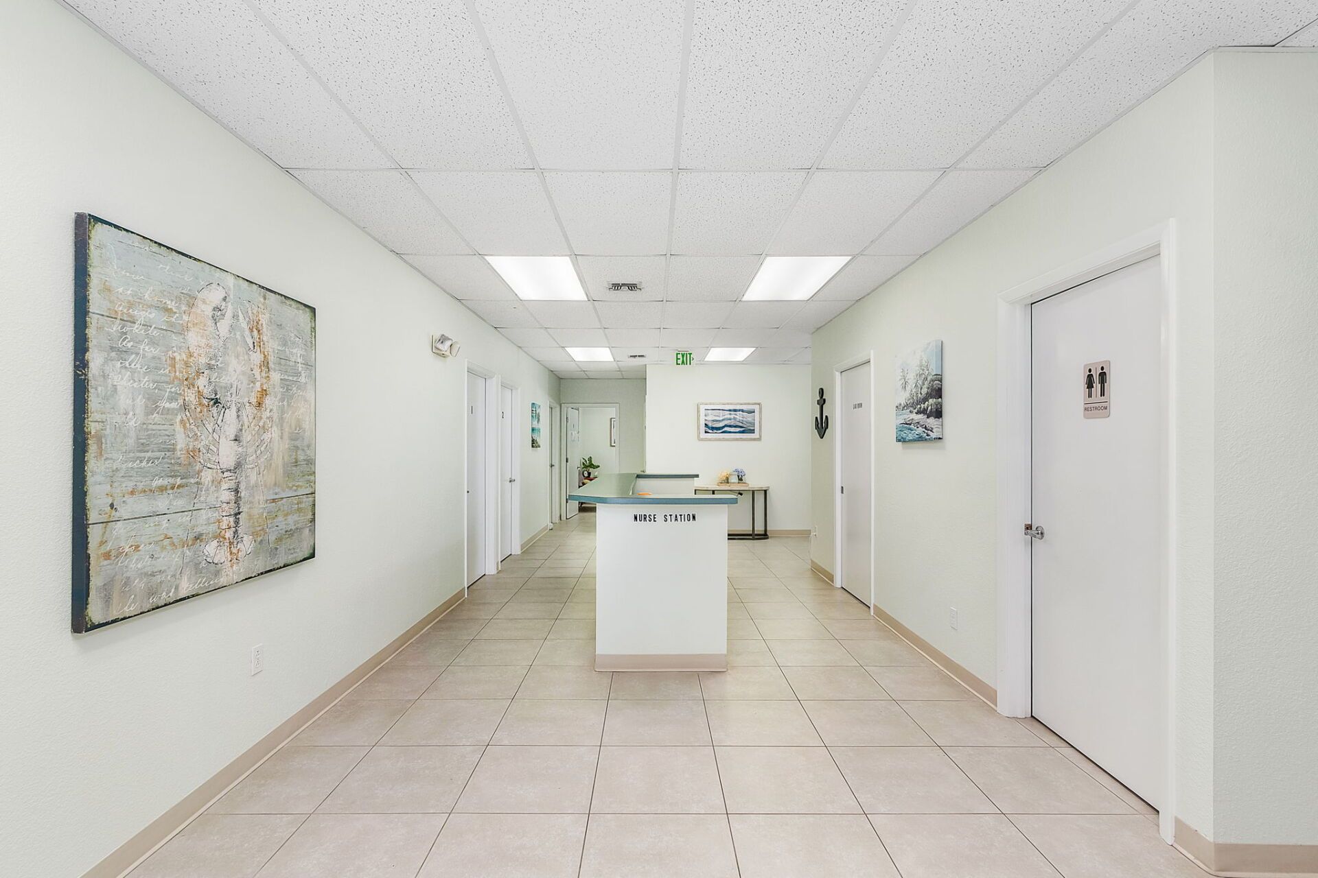 Bright, empty office hallway with white walls, tiled floor, central cabinet, and framed artwork.