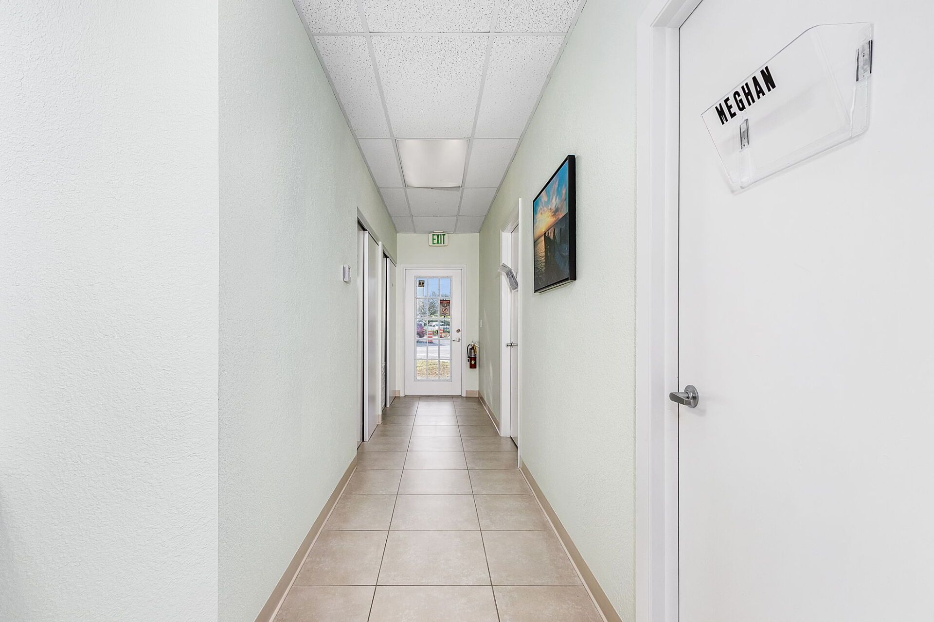 Bright white hallway with closed doors, tiled floor, and framed wall art leading to a window at the end.