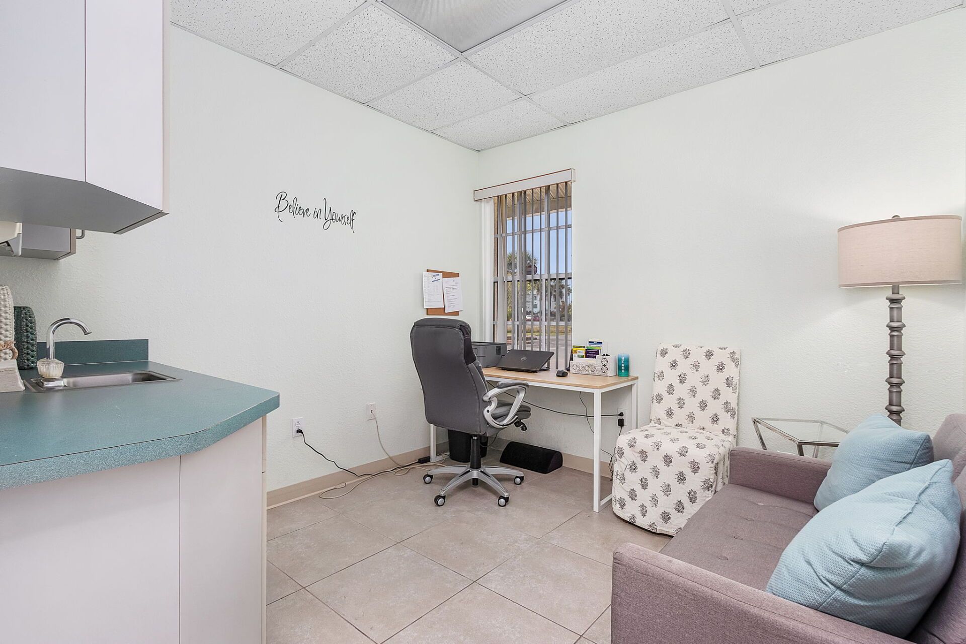 Small home office with a desk, rolling chair, gray sofa, floor lamp, and turquoise kitchen counter.