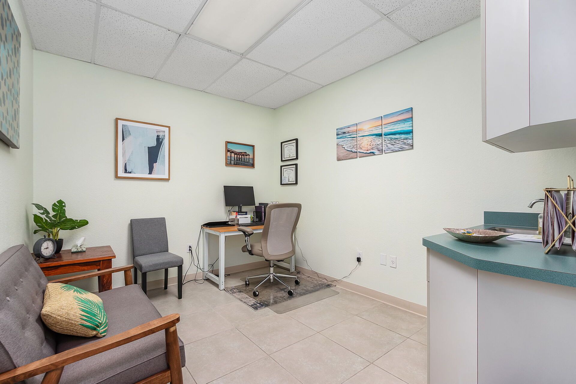 Small office with desk, chair, computer, wall art, and a gray sofa in a bright room