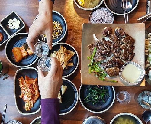 Two people toasting shots over a Korean meal, featuring various side dishes and grilled mushrooms.