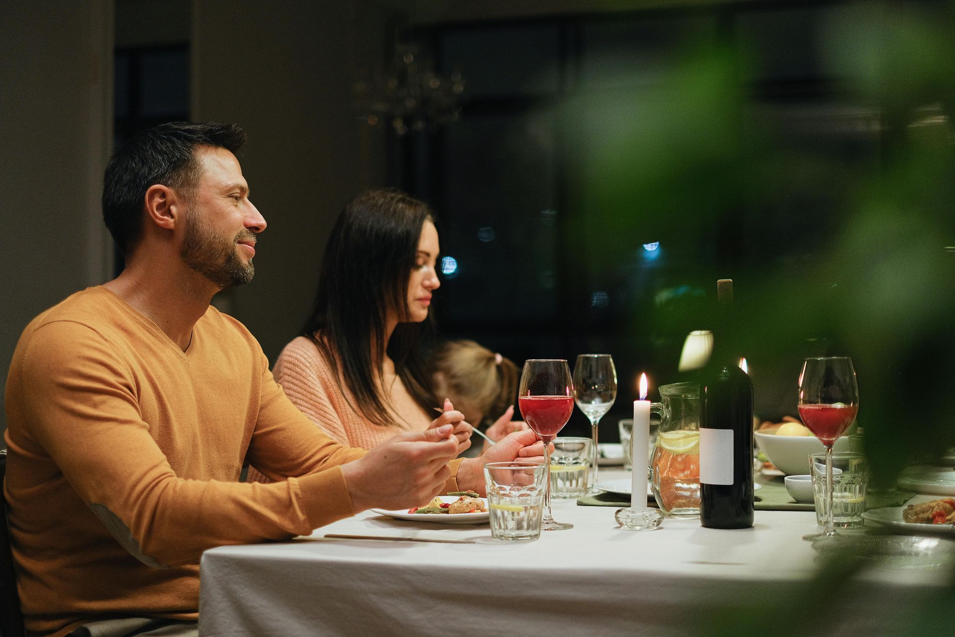 Couple at a dinner table, lit by candles and wine glasses, with food and a bottle of wine.
