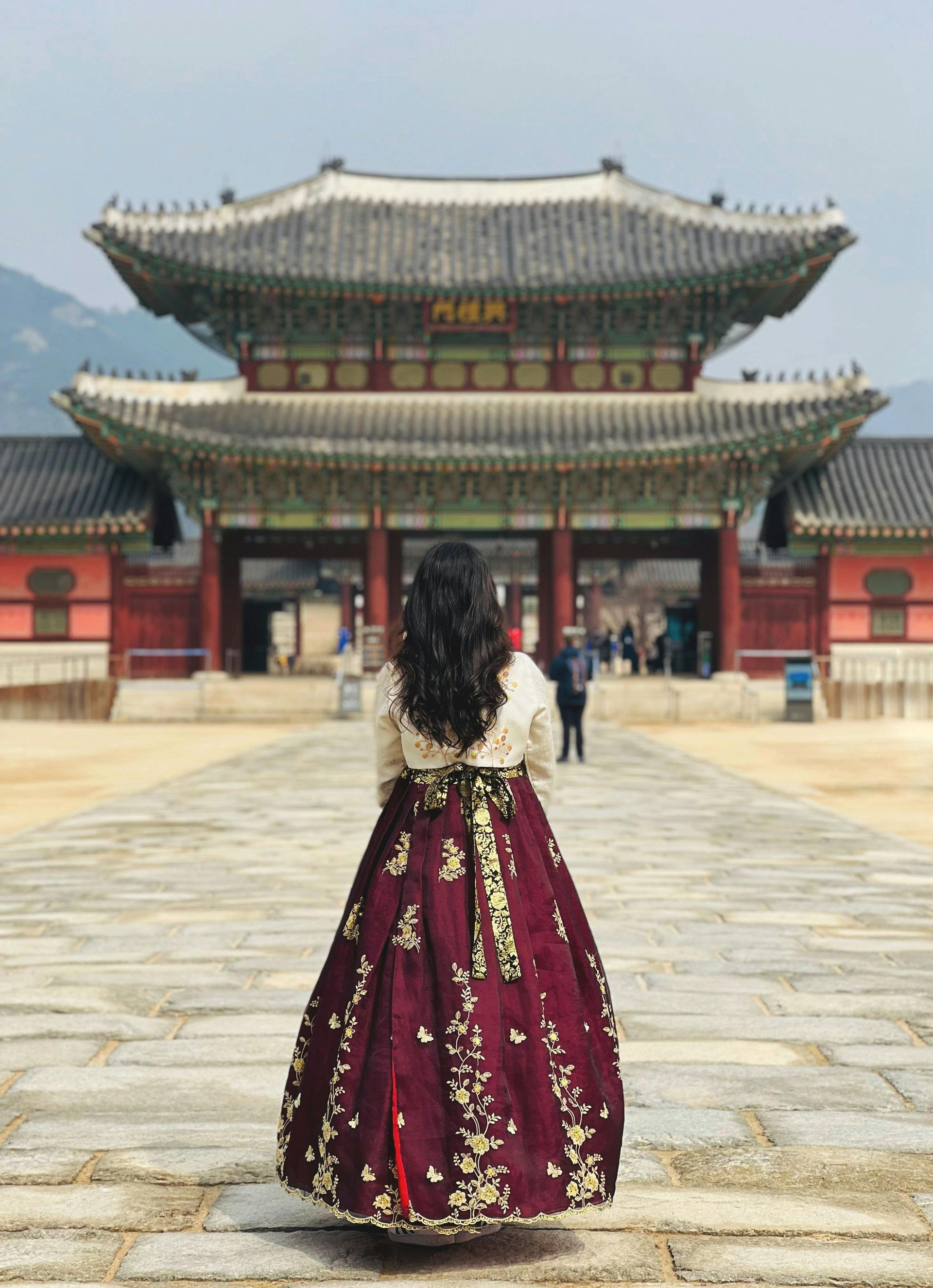 Woman in hanbok facing a Korean palace entrance, maroon skirt, long black hair.