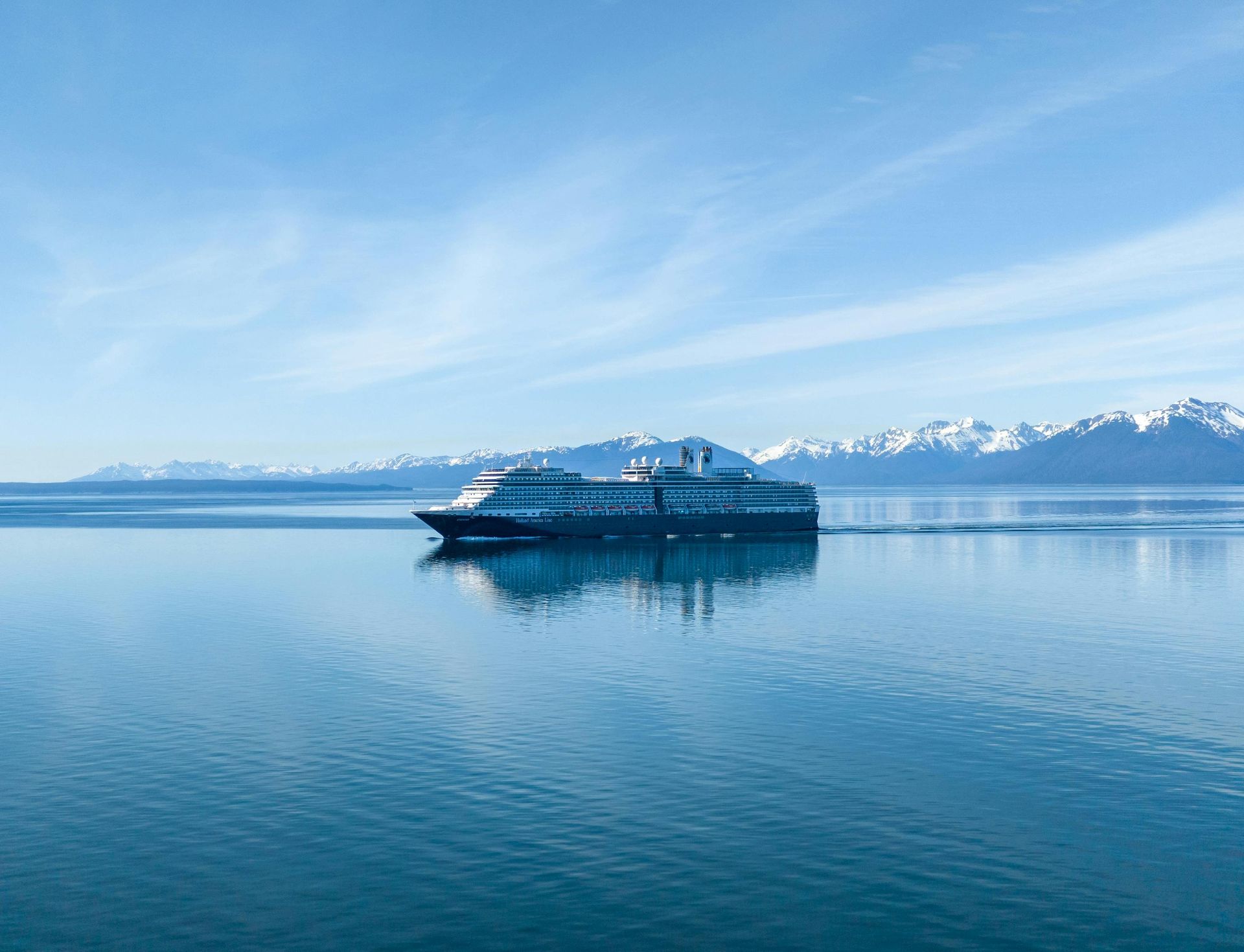 Cruise ship sails on calm water, mountains in background under a blue sky.