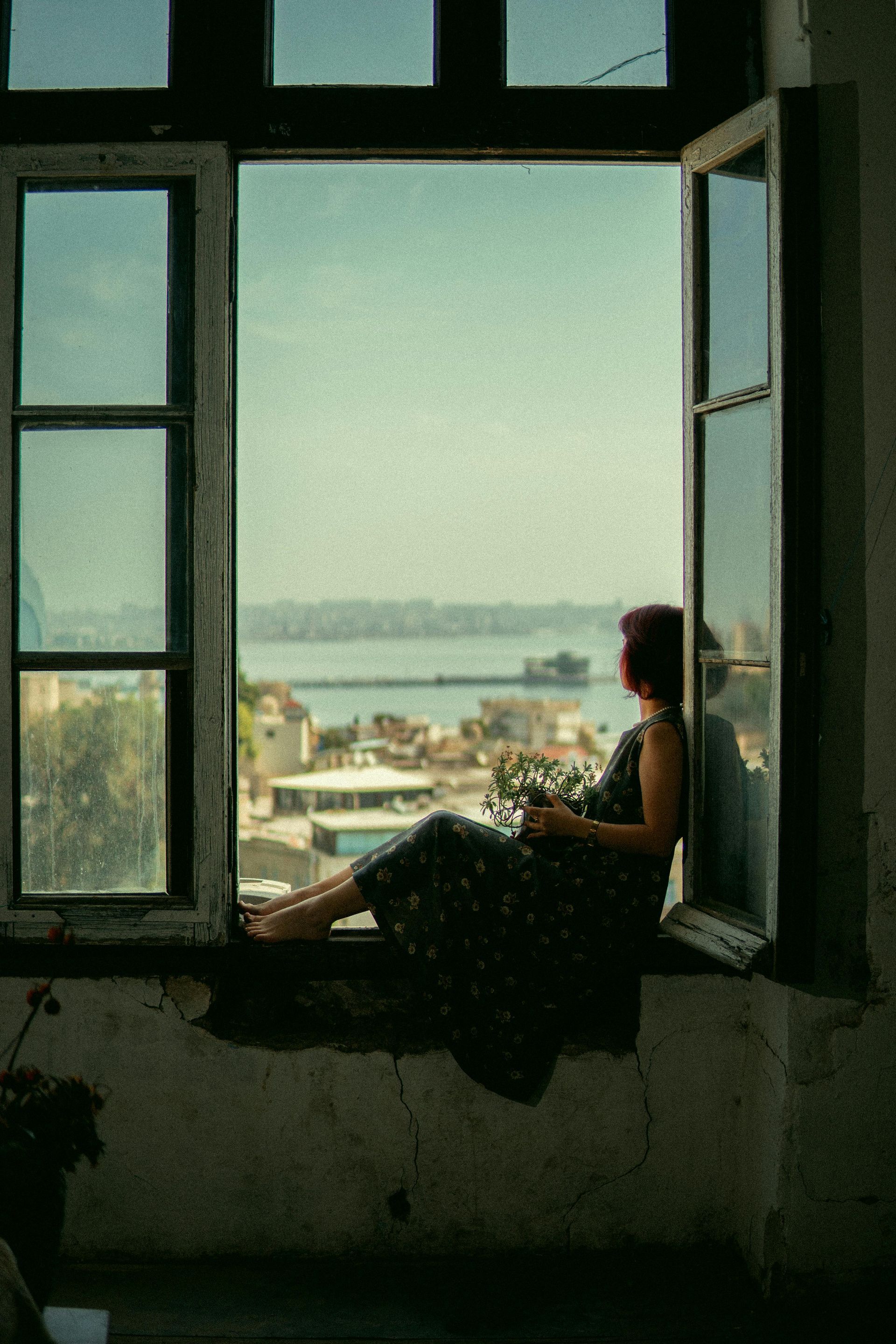 Woman sitting in open window, overlooking a city and water. Holding flowers, relaxed.