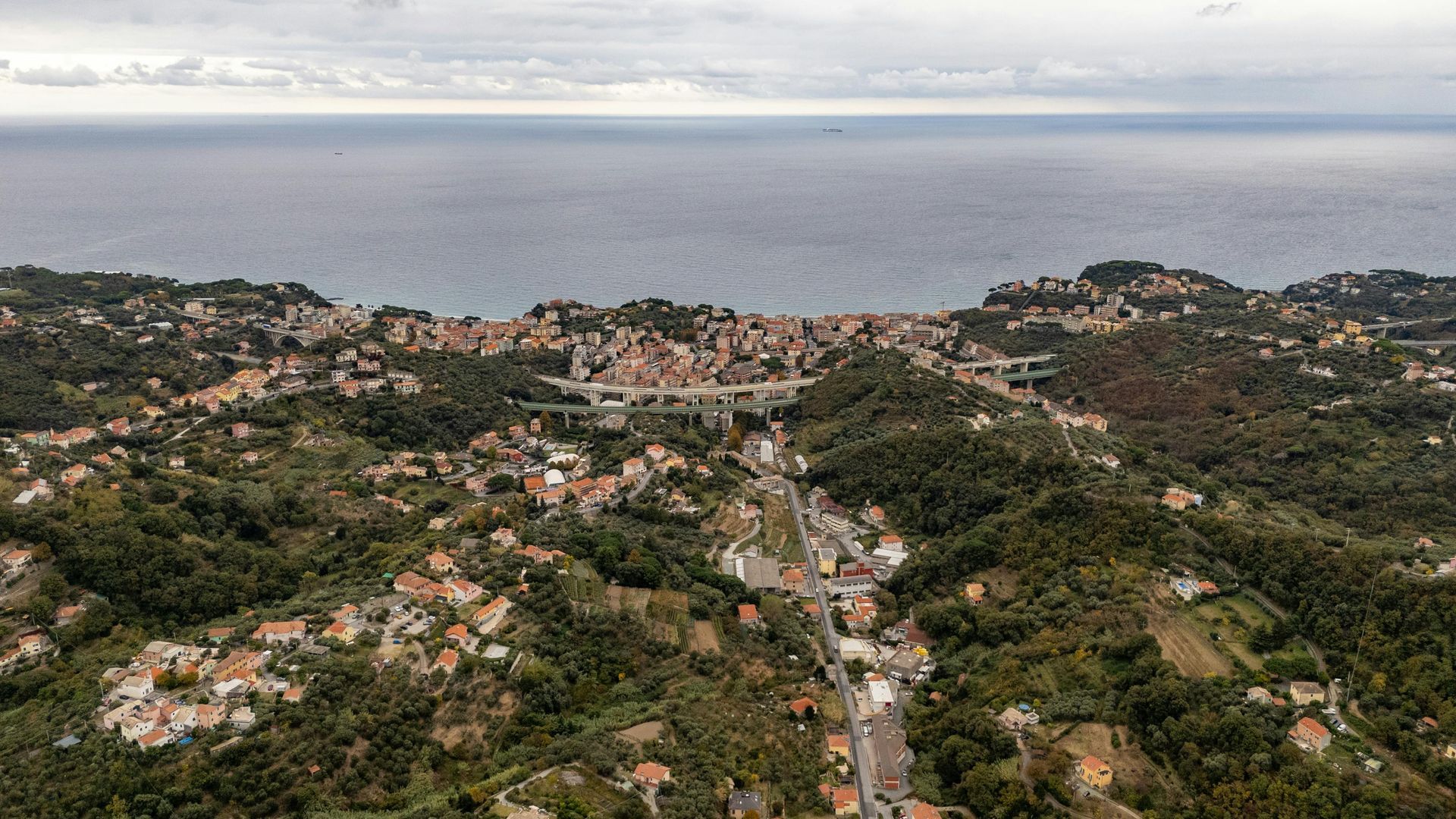 Coastal town nestled in green hills, overlooking the sea under a cloudy sky.