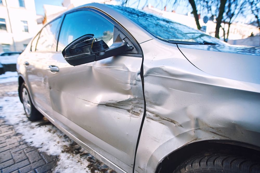 A silver car with a damaged side door is parked in the snow.