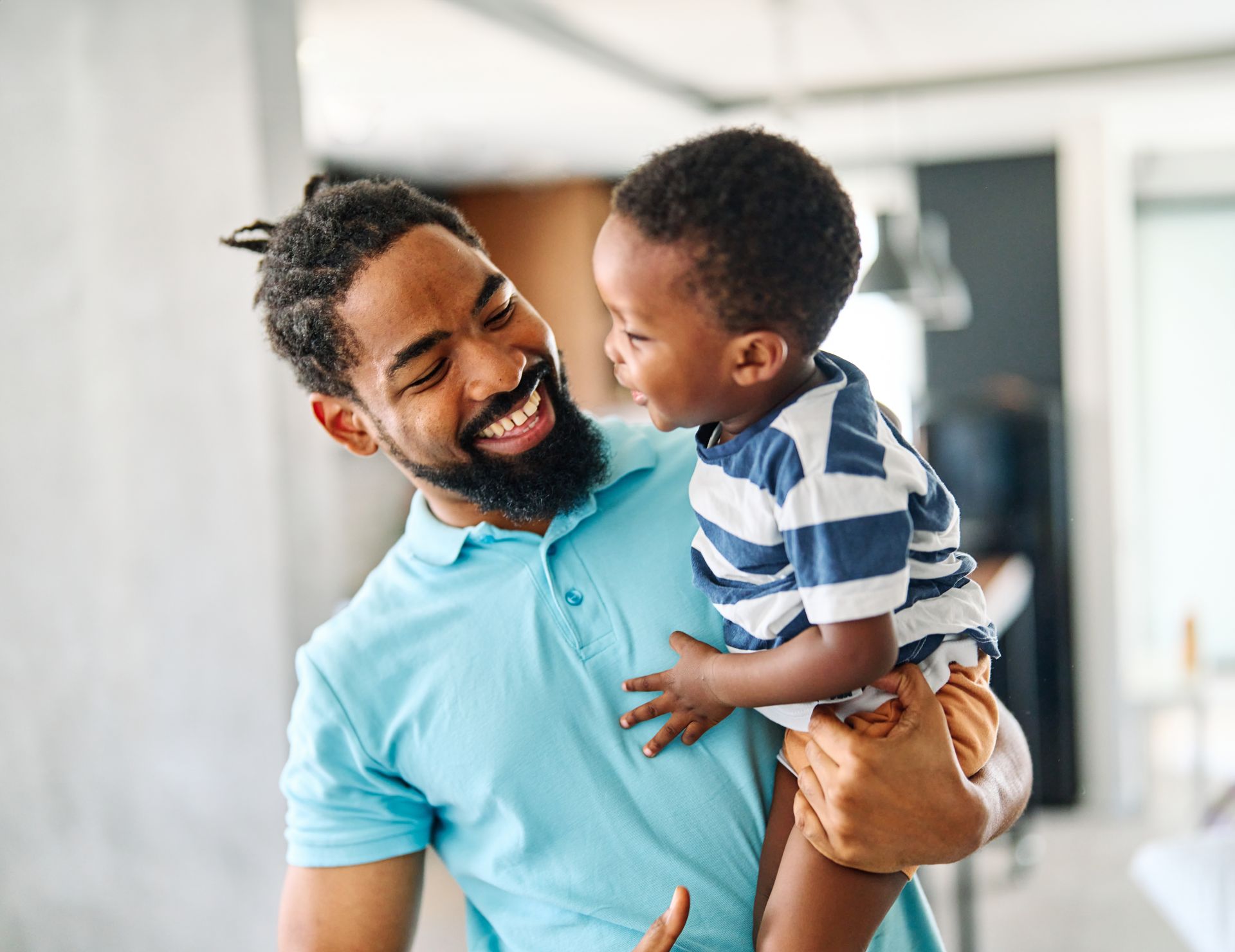 A smiling person wearing a blue polo shirt holds a young child in a striped shirt, both looking at each other warmly.