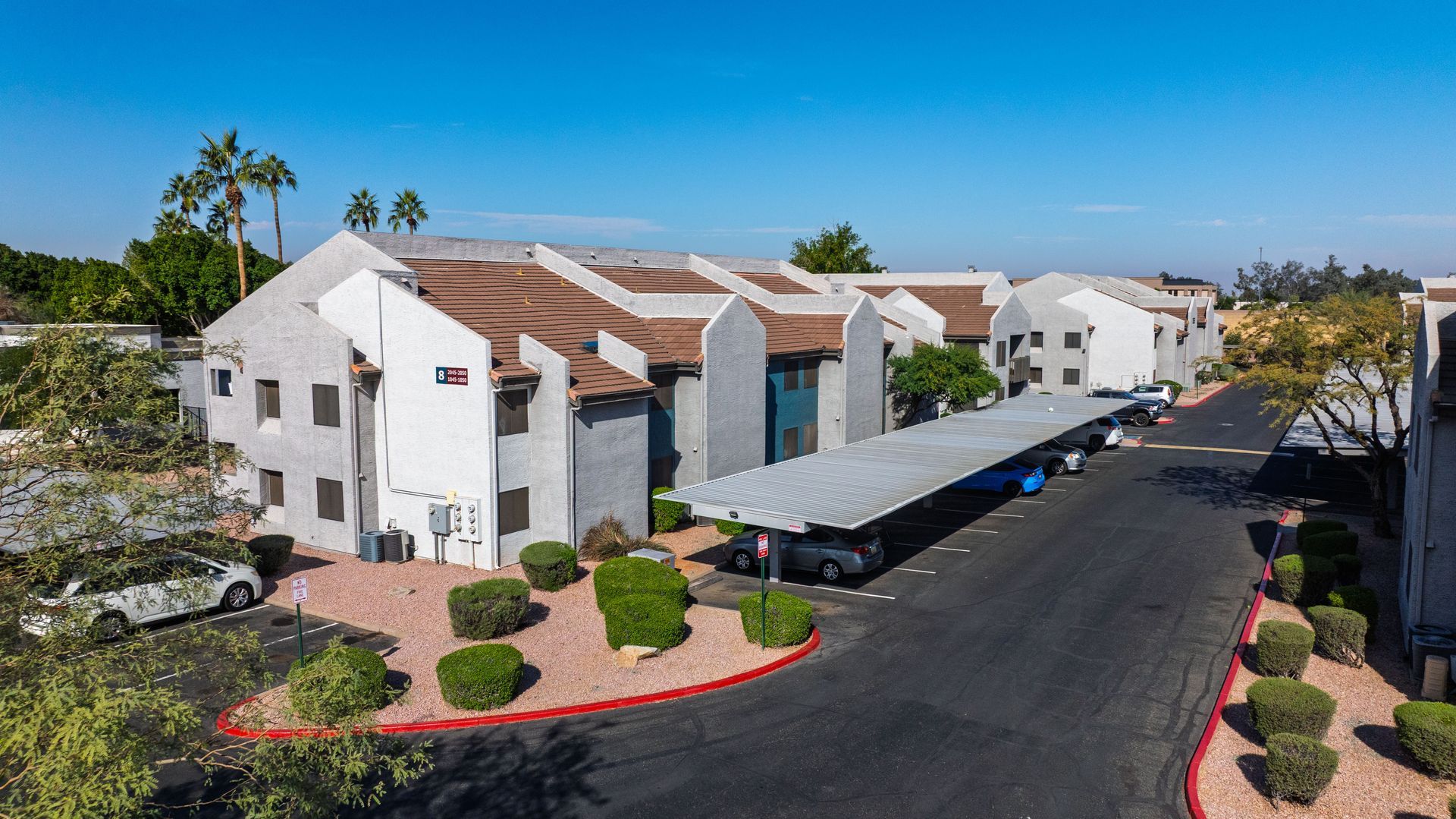 Apartment complex with a long covered parking area, palm trees, and blue sky at the Maddox.