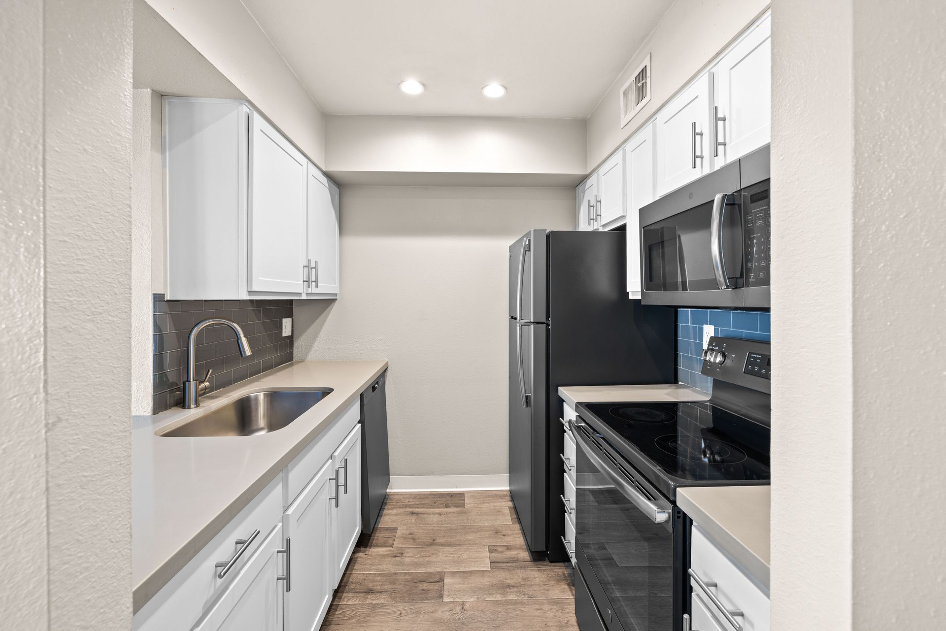 A galley kitchen with white cabinets, stainless steel sink, black appliances, and light wood flooring at the Maddox.