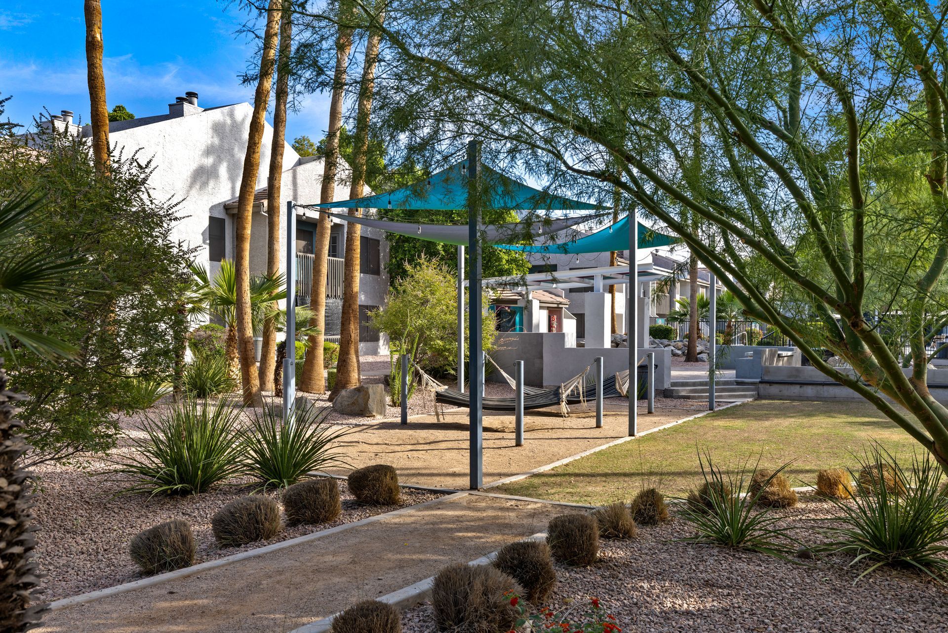 A gravel path leads to a shaded structure with turquoise canopies, surrounded by landscaping, trees, and a building.