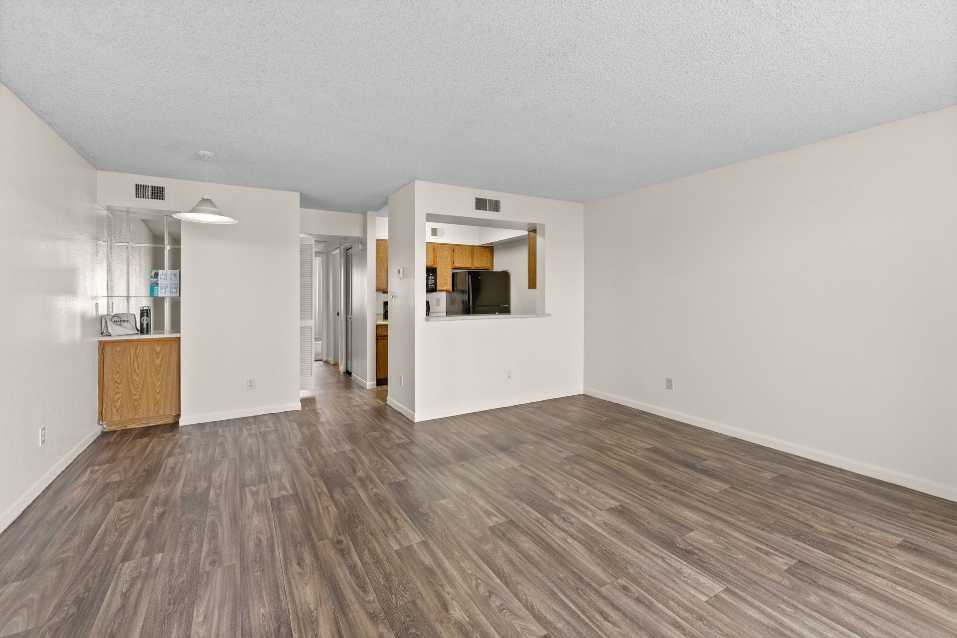 Empty living room with wood-look flooring, opening to kitchen, and hallway. White walls and ceiling.