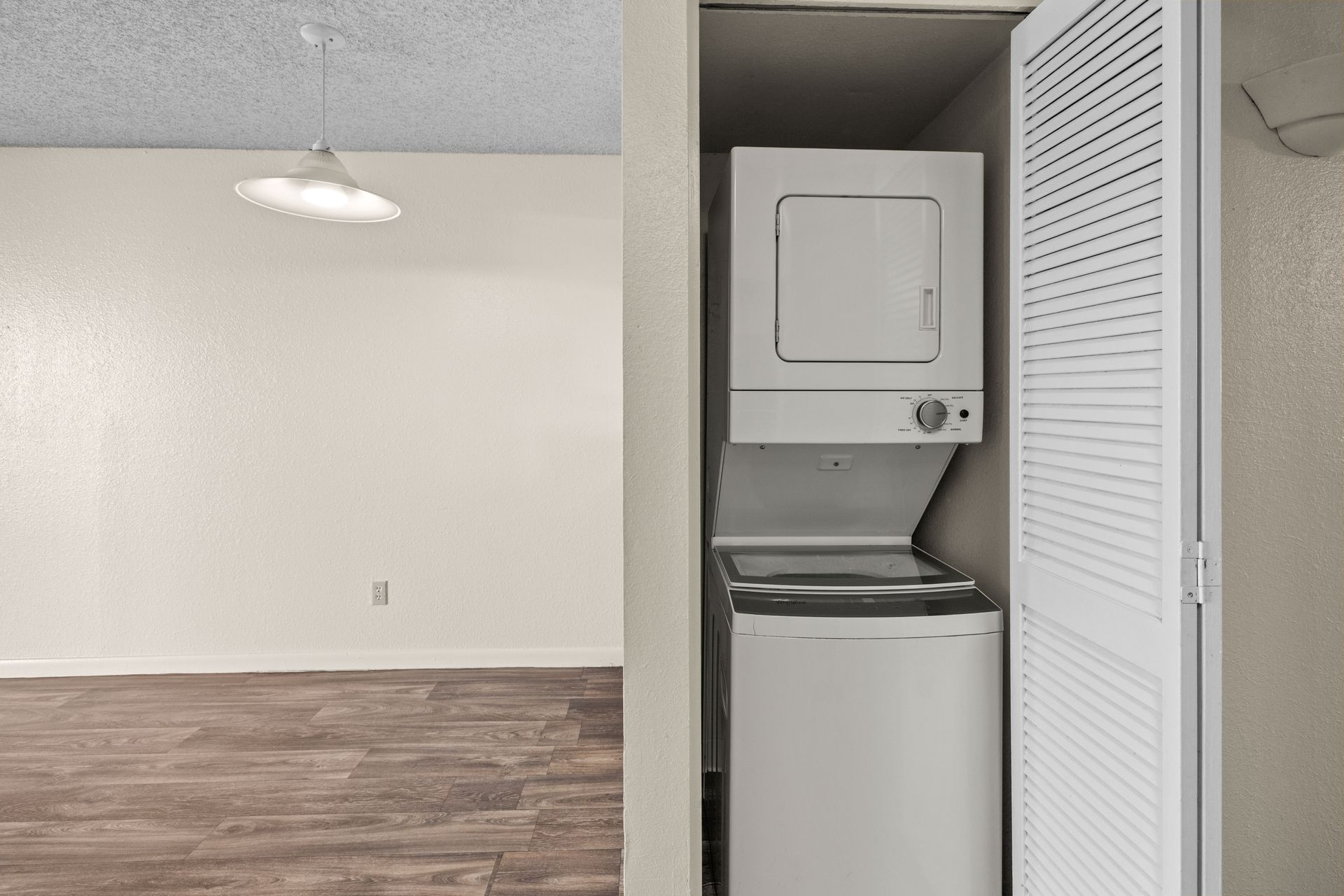Stacked white washer and dryer in a laundry closet, next to an open, louvered door. Room with hardwood floor visible.