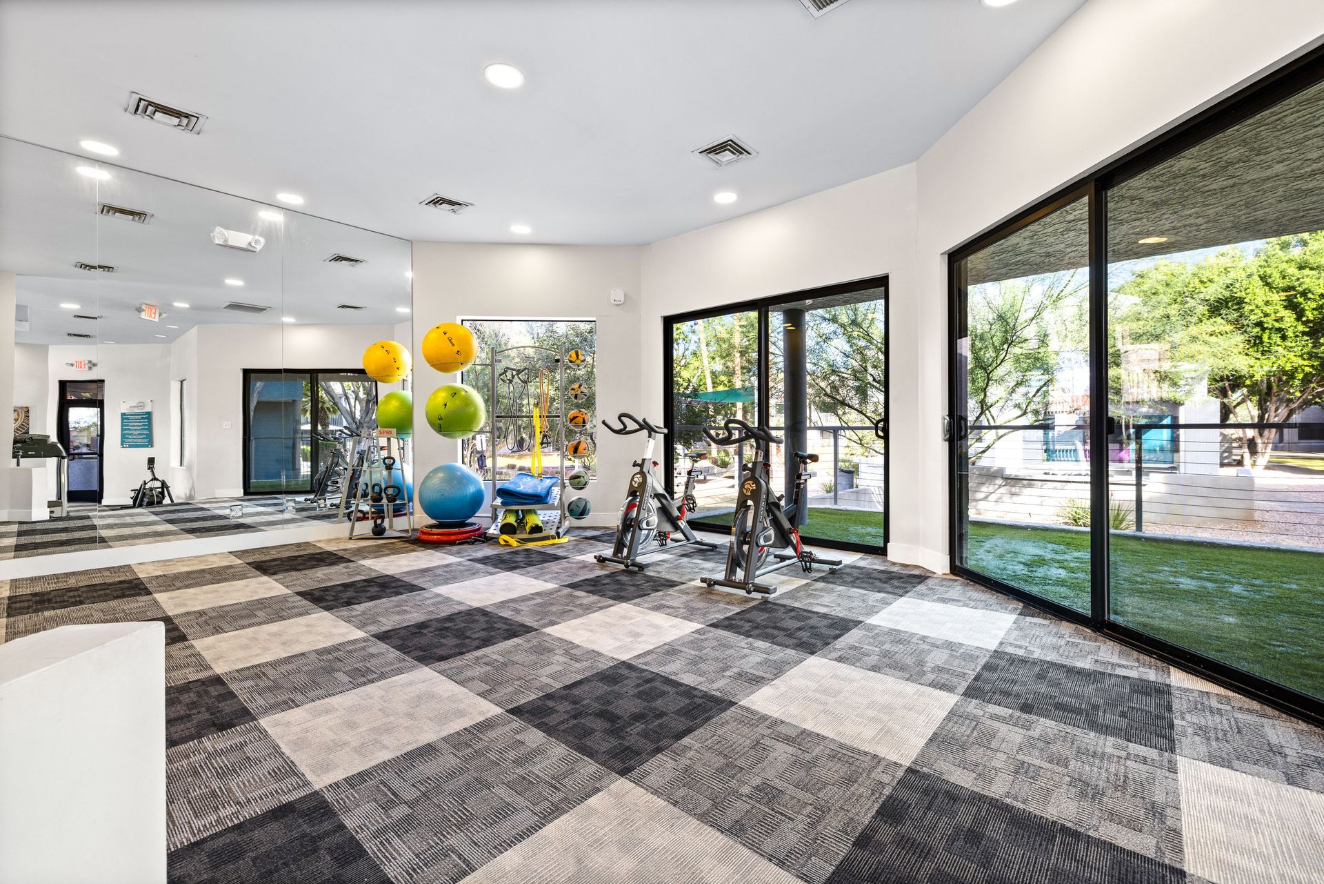 Gym with checkered floor, exercise equipment, mirrored wall, and large windows overlooking outdoor space.