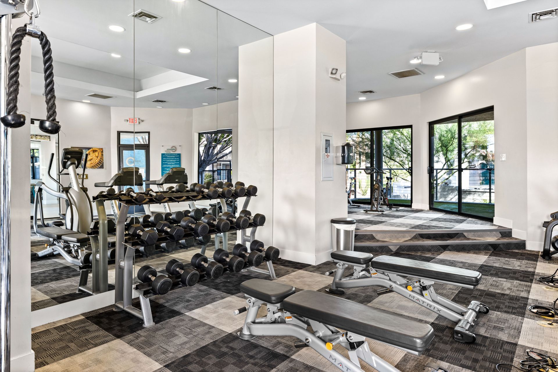 Gym with weights, benches, and cardio equipment. Mirrored wall reflects equipment, black and gray checkered floor, natural light from windows.