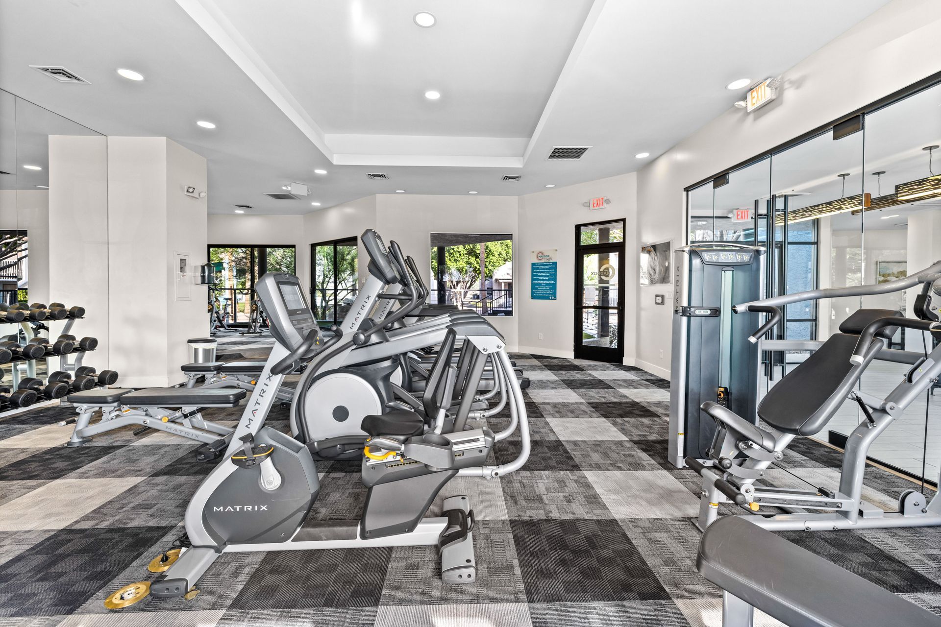 Gym with exercise equipment, including elliptical machines and weights, on a patterned floor.