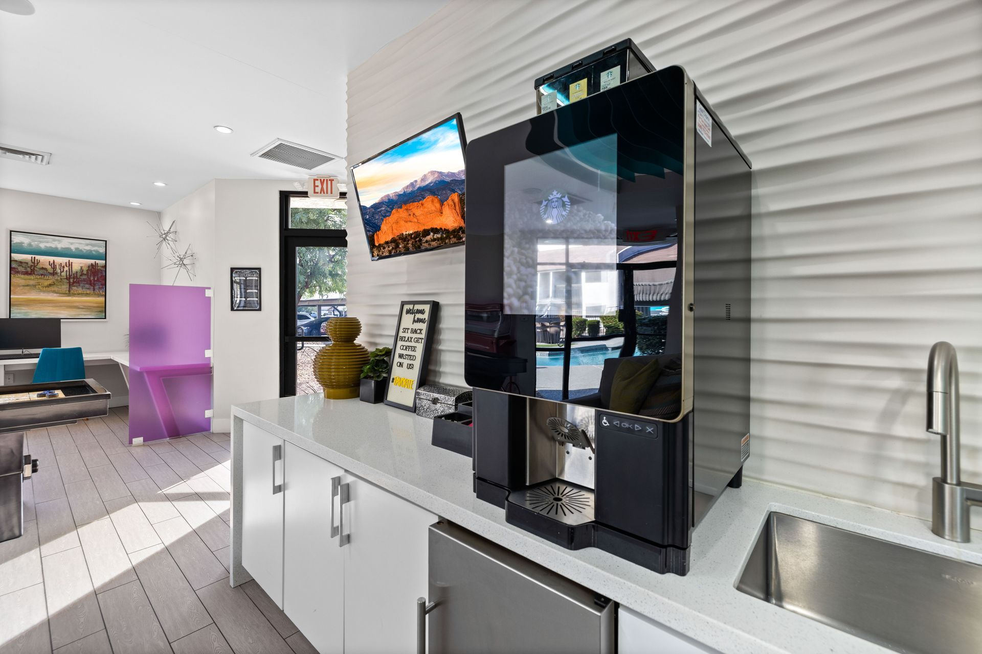 Coffee machine on white countertop in a bright room with a tv and sink.