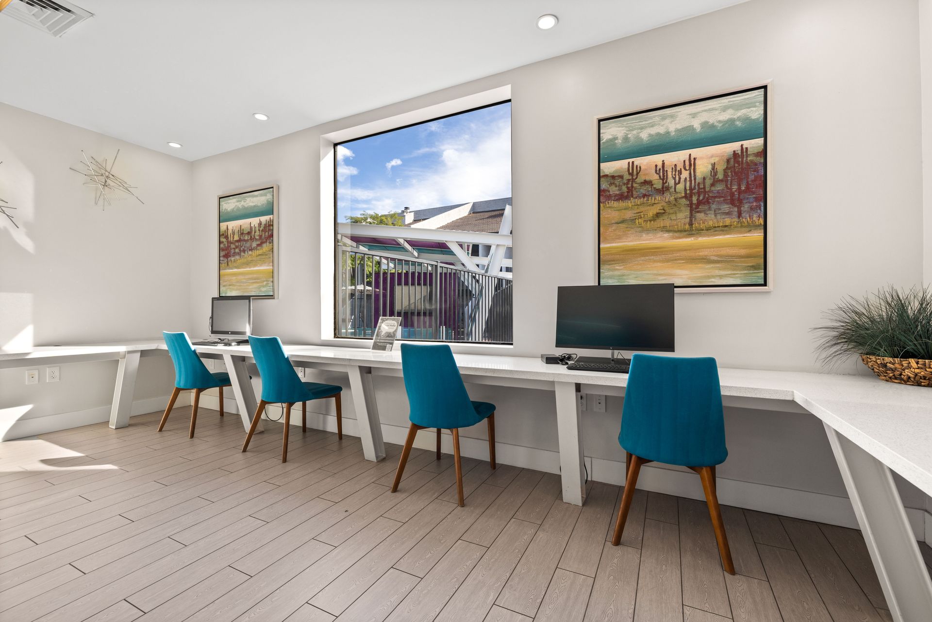 Bright office space with long white desks, turquoise chairs, computers, and artwork. A large window overlooks an outdoor space.