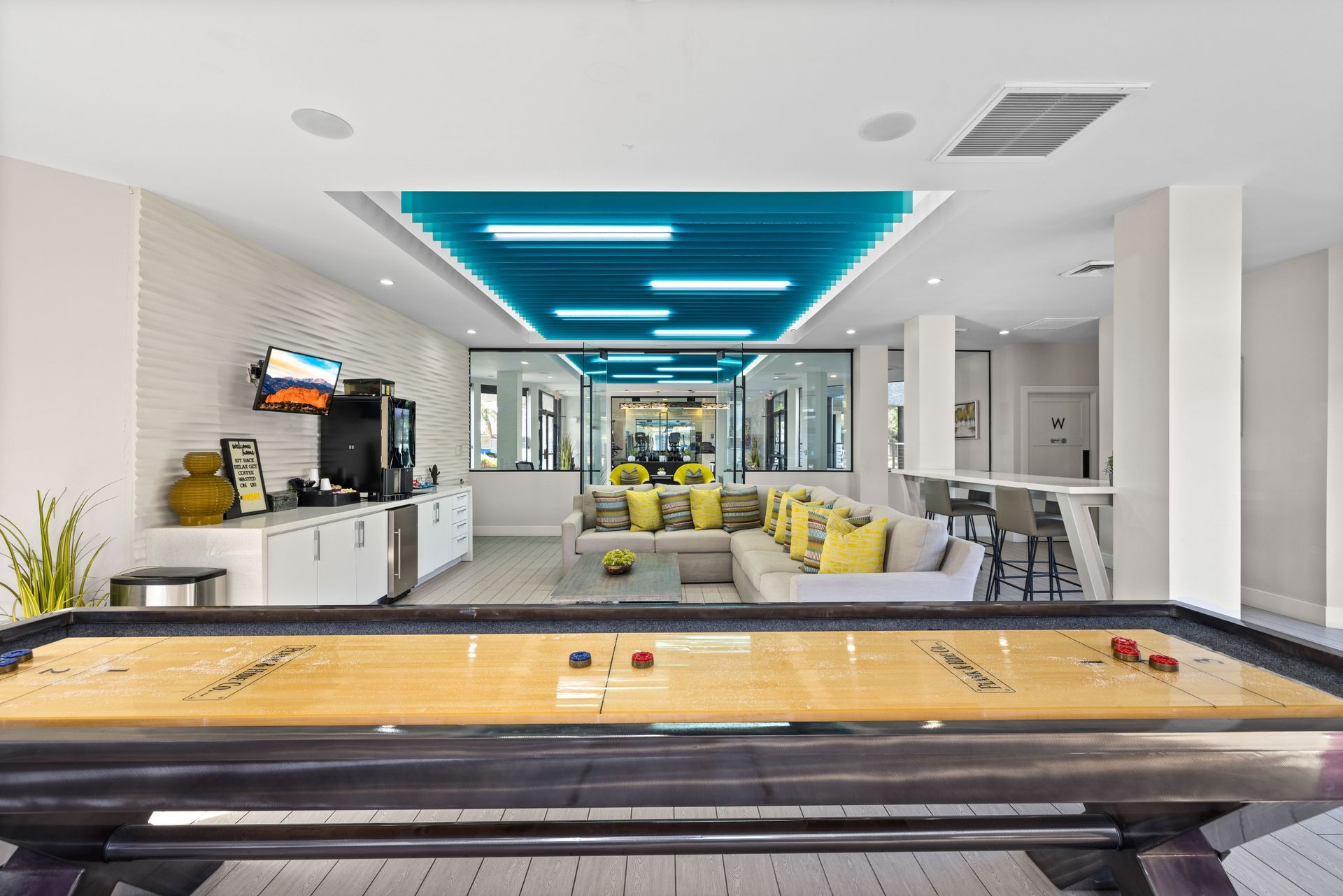 A bright, modern recreational room with a shuffleboard table, lounge area, and a coffee station.