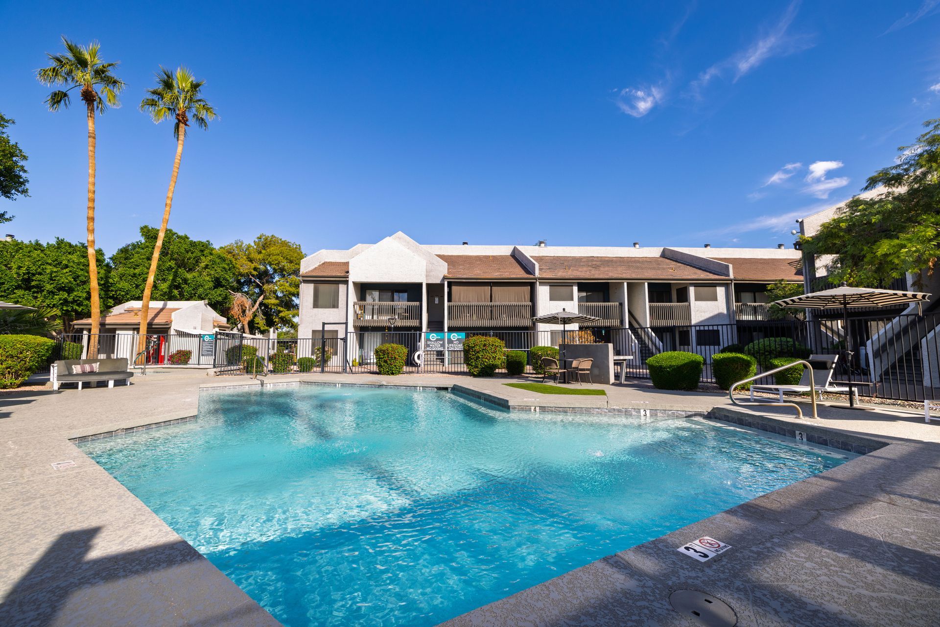 Pool in front of a two-story apartment building under a blue sky, palm trees at the Maddox.