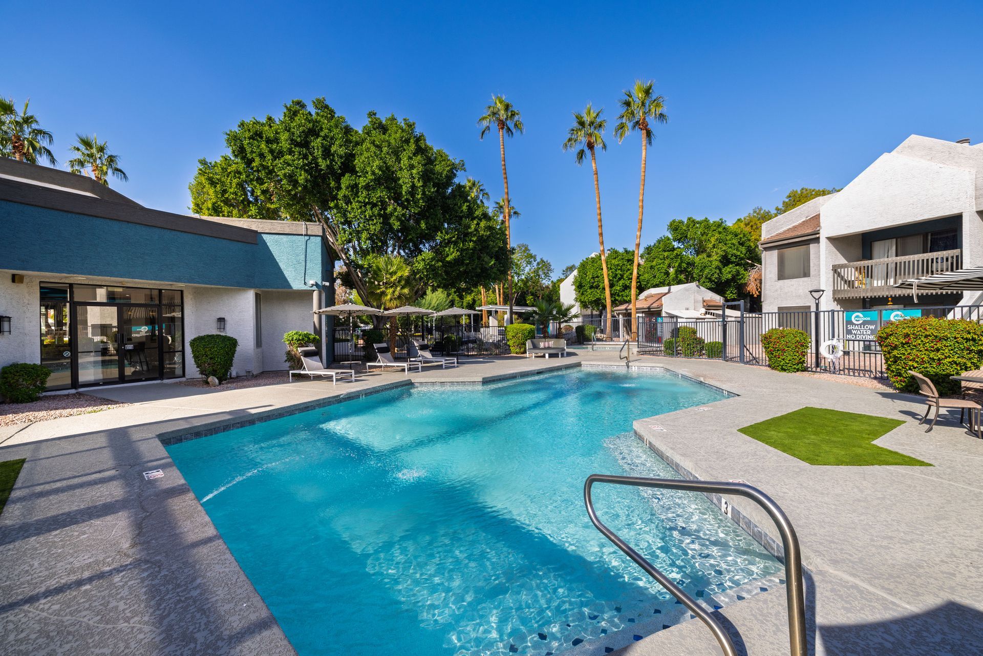 Pool with turquoise water surrounded by concrete, palm trees, and two-story buildings on a sunny day.