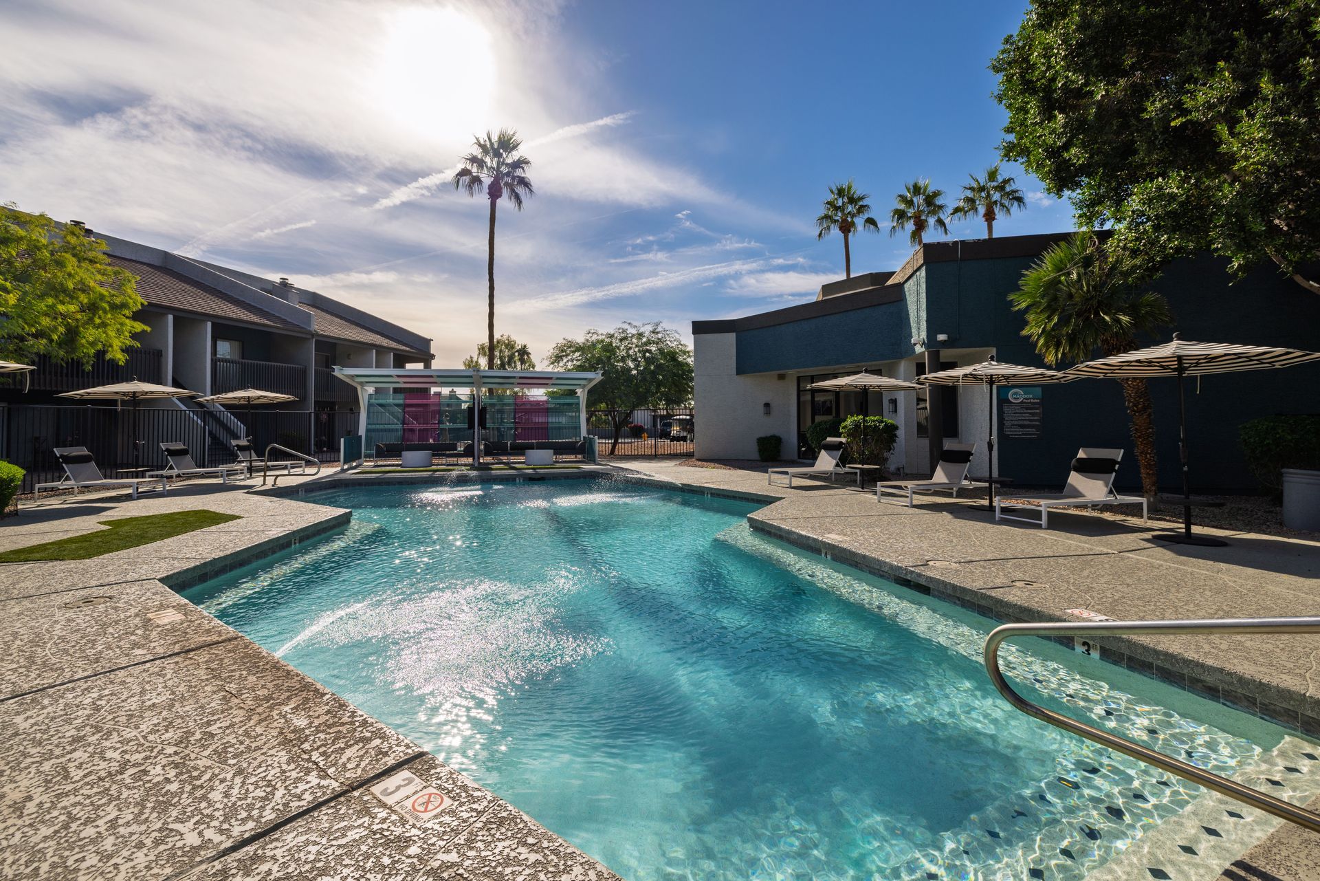 Pool with clear blue water surrounded by lounge chairs, palm trees, and buildings under a sunny sky.